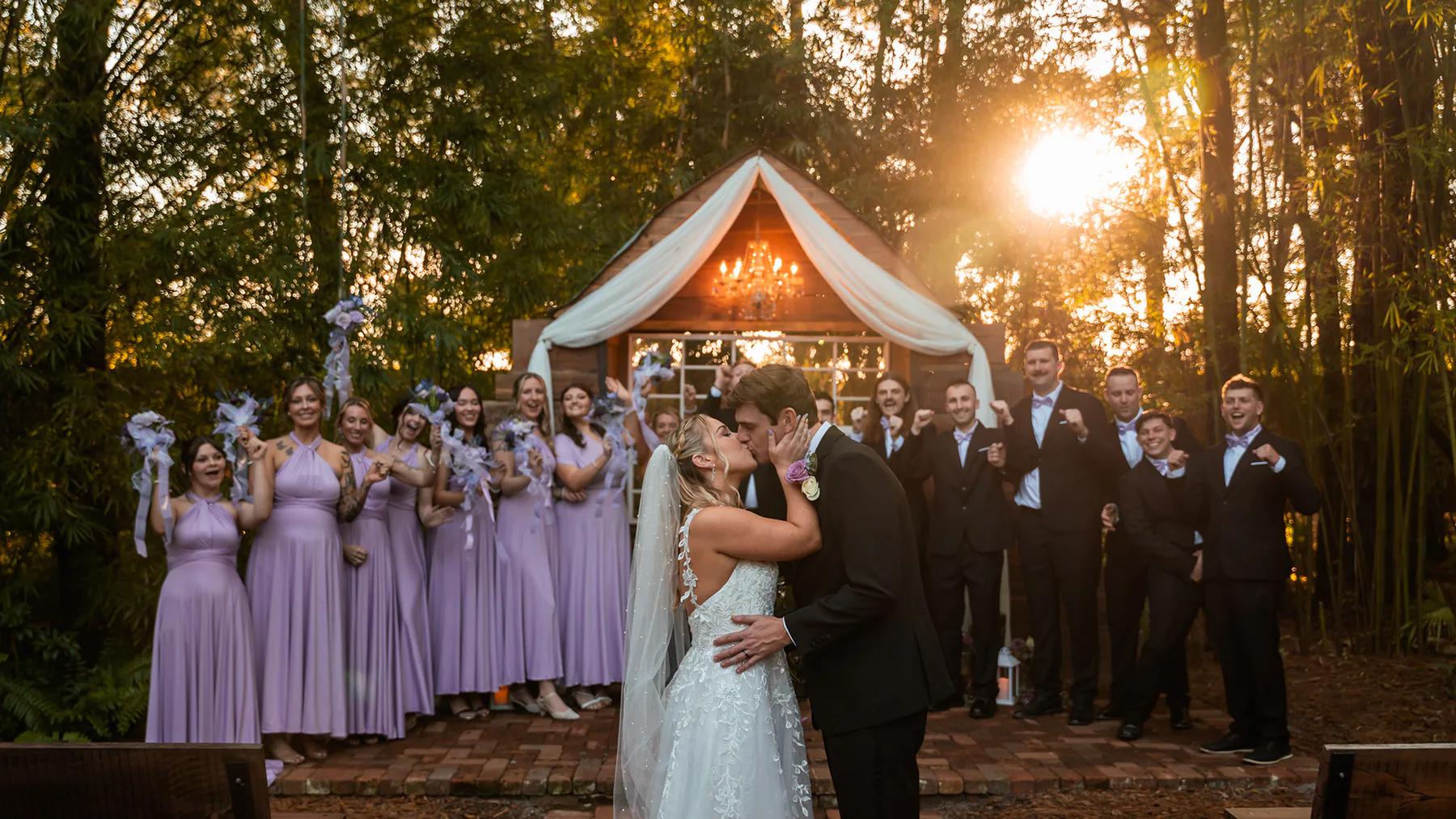 Wedding party applauding as the newlyweds kiss beneath a draped outdoor arch.