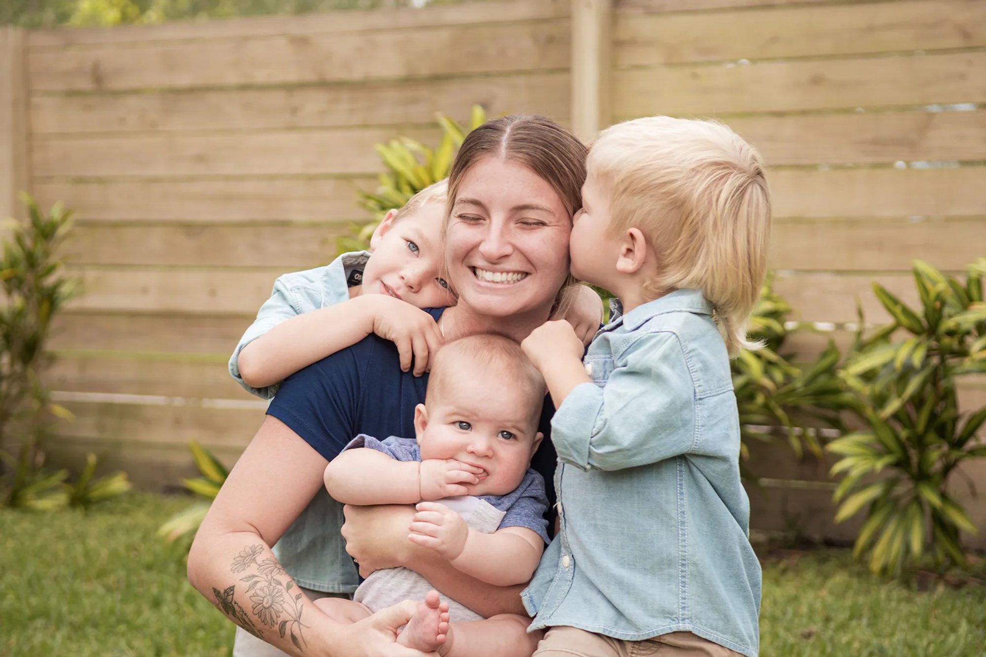 Mother holding a baby while two young boys kiss her cheeks.