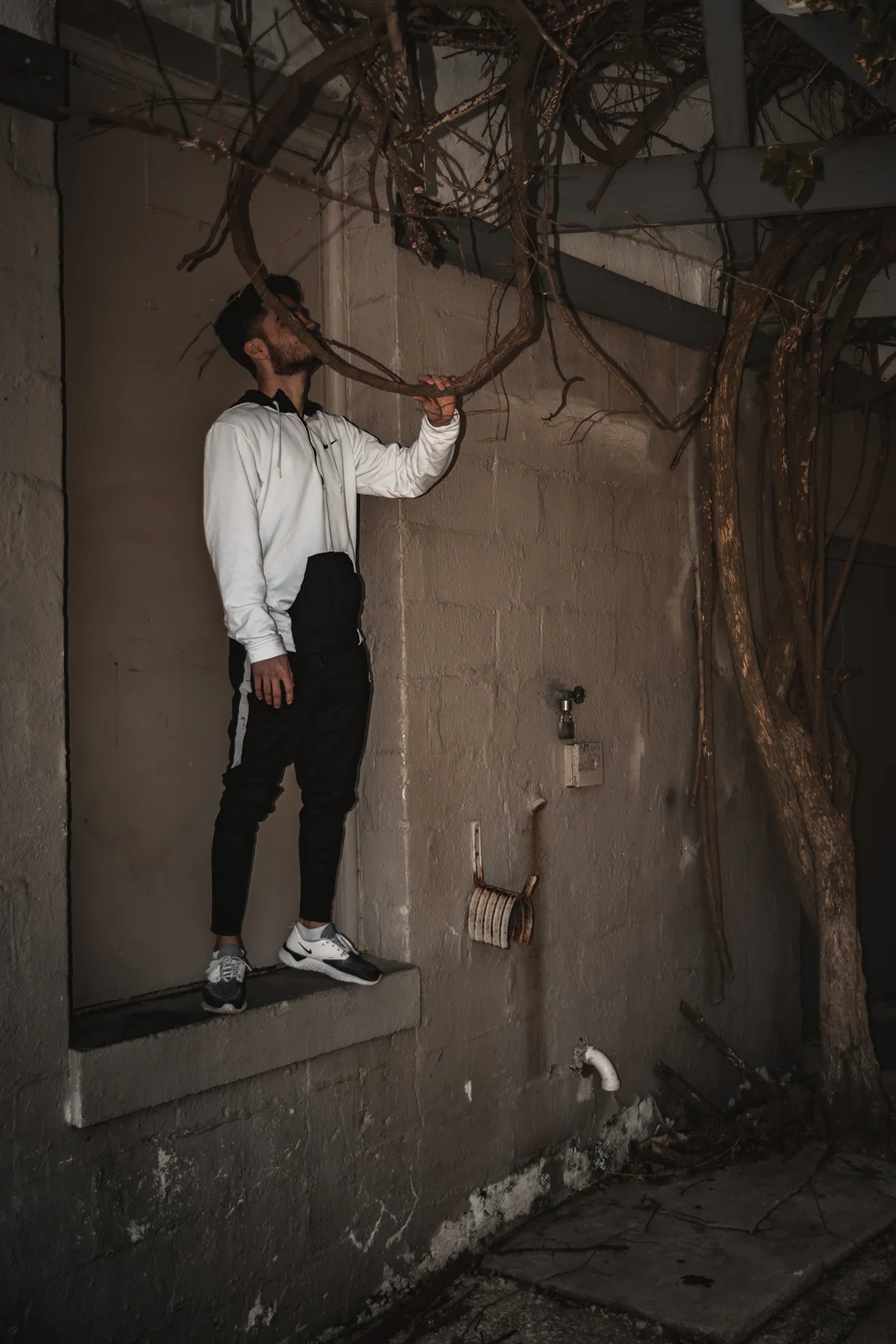 Man in a white hoodie posing beside a bare tree and weathered wall.