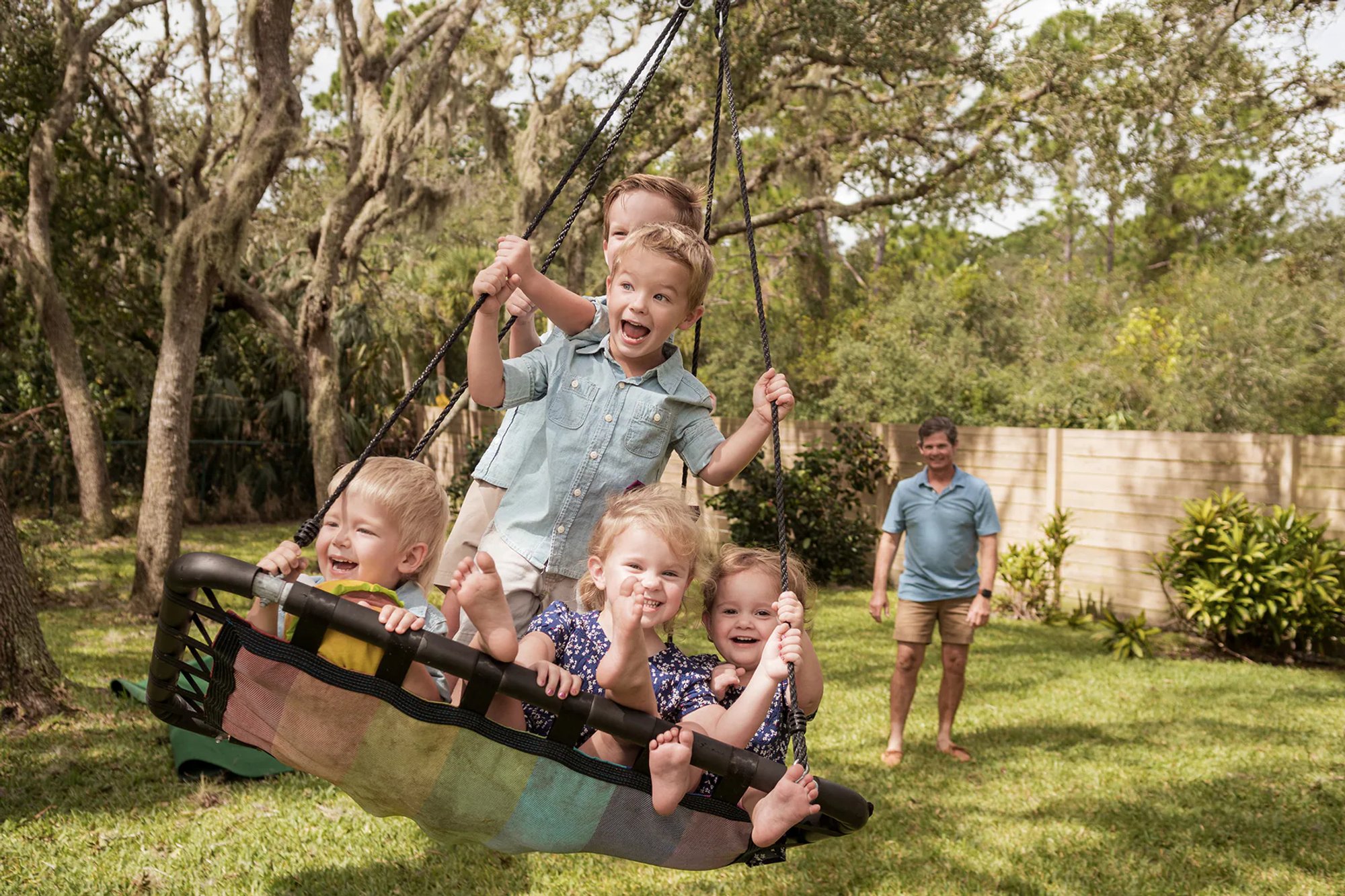 Children smiling on a large swing while an adult stands behind them.