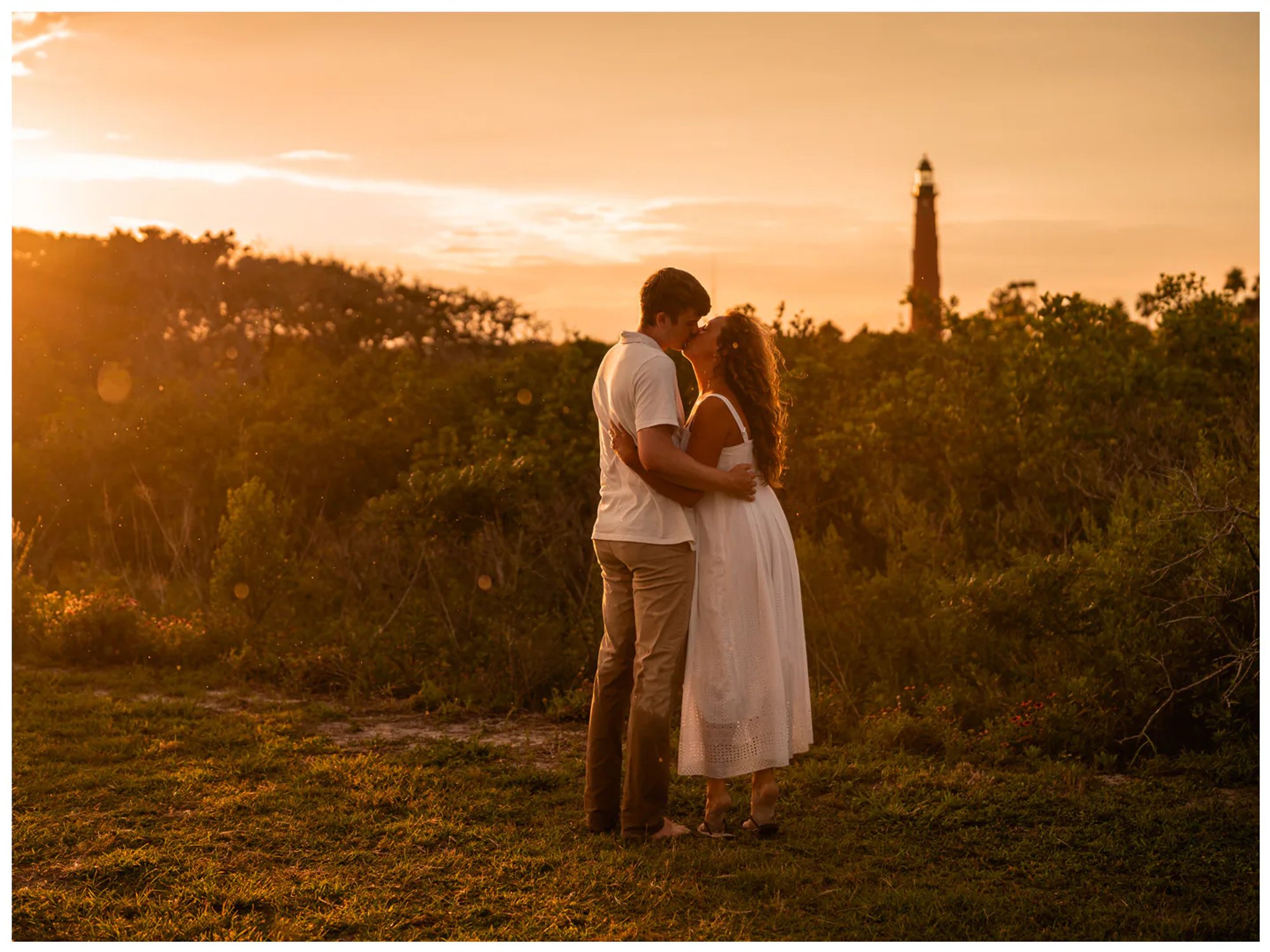 Couple embracing near coastal dunes with a lighthouse in the distance.