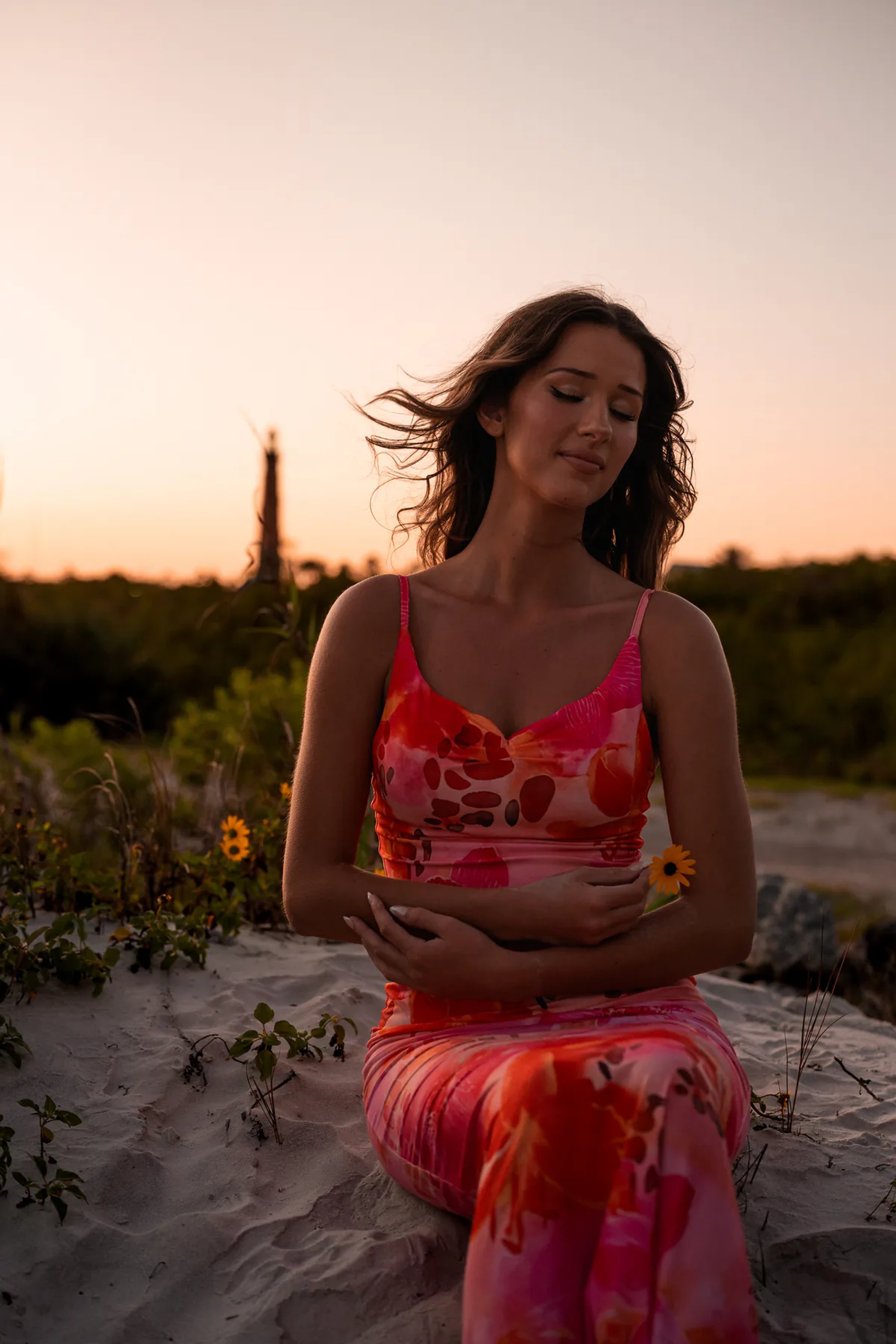Young woman in a coral patterned dress seated among coastal plants.