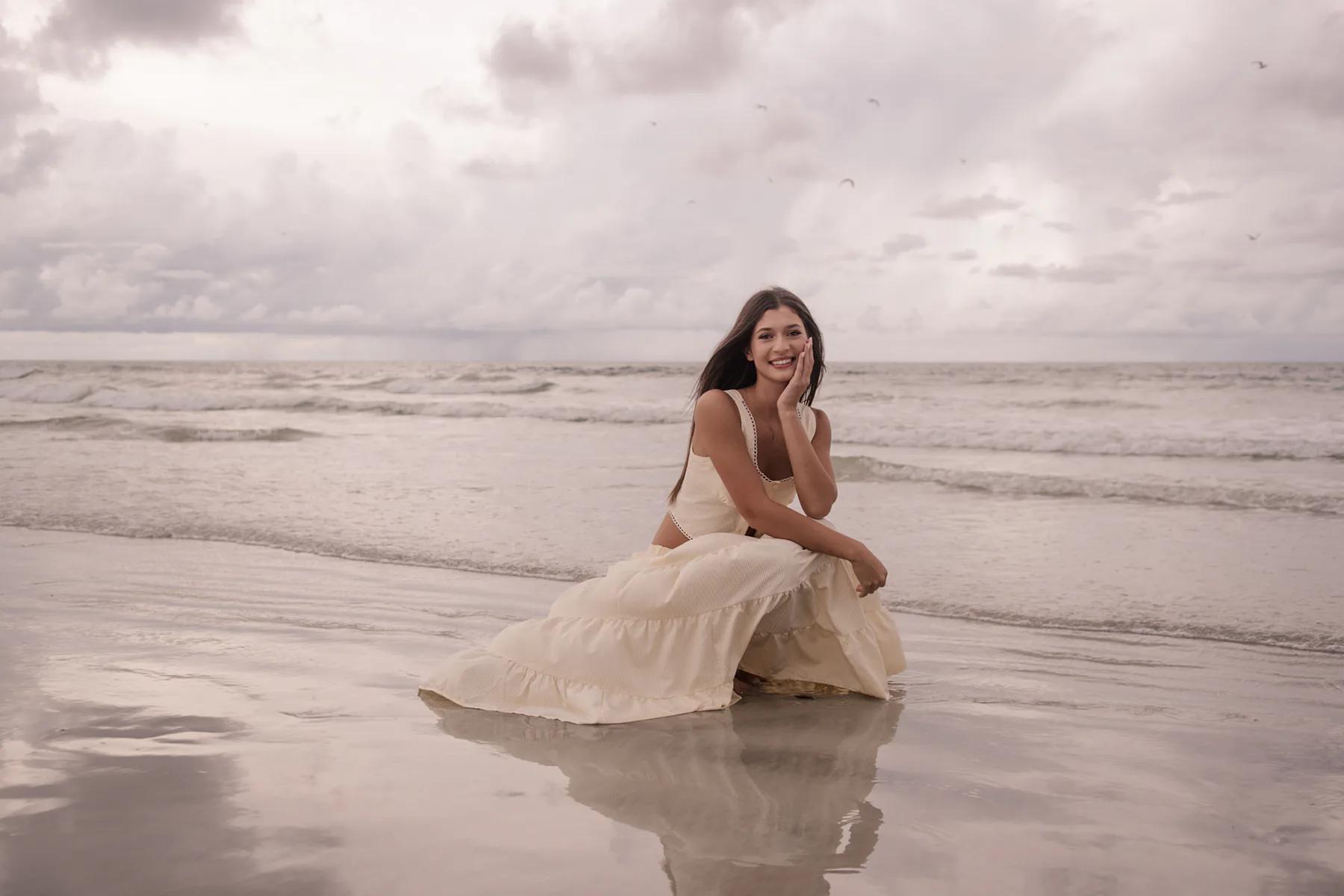 Senior portraits on Ponce Inlet Beach with Mia sitting on the shoreline in a cream dress, natural light photography in Daytona Beach FL.