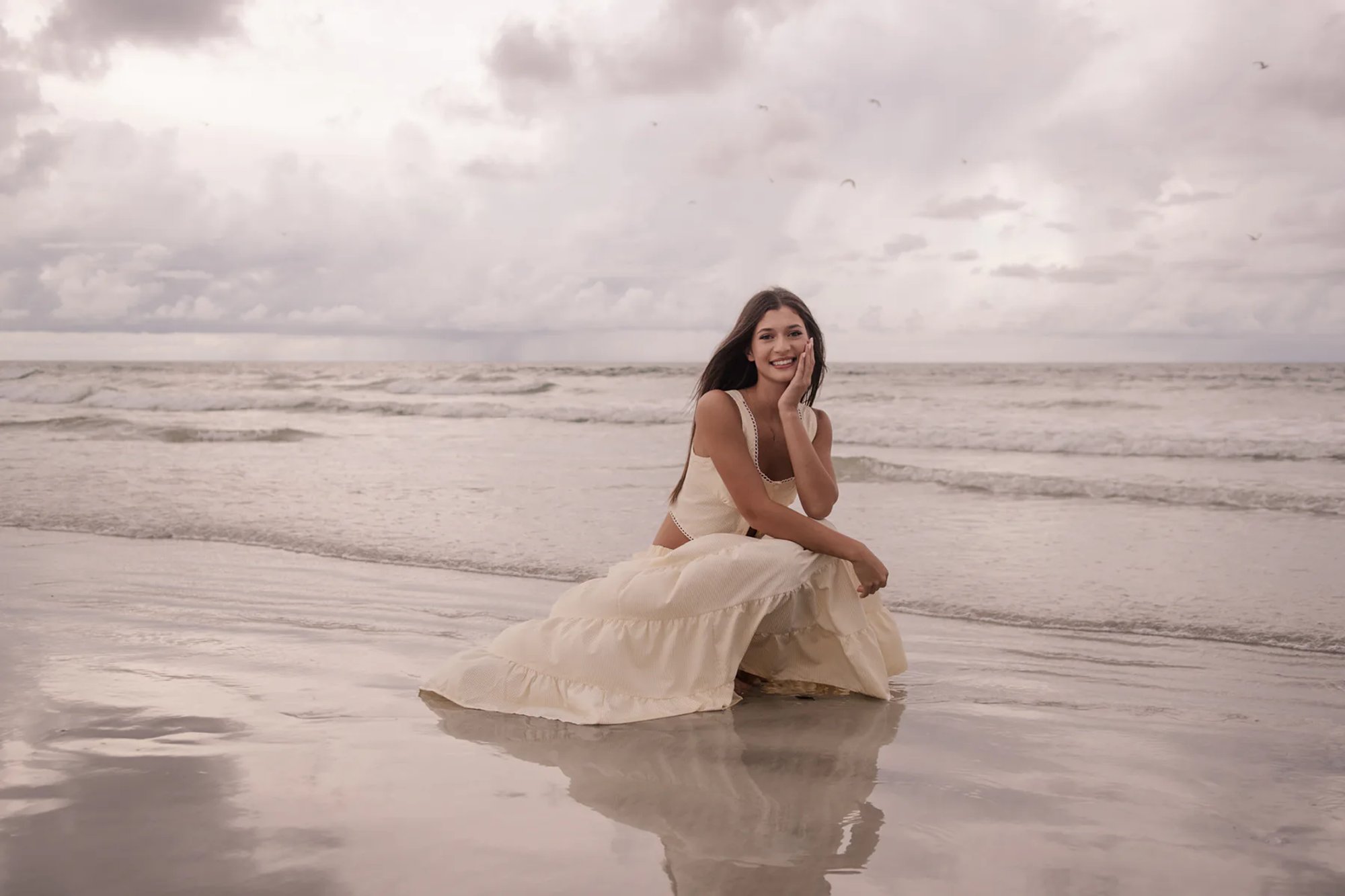 Senior portraits at Ponce Inlet Beach, Mia sitting at the shoreline in a cream dress with soft reflection in the water.