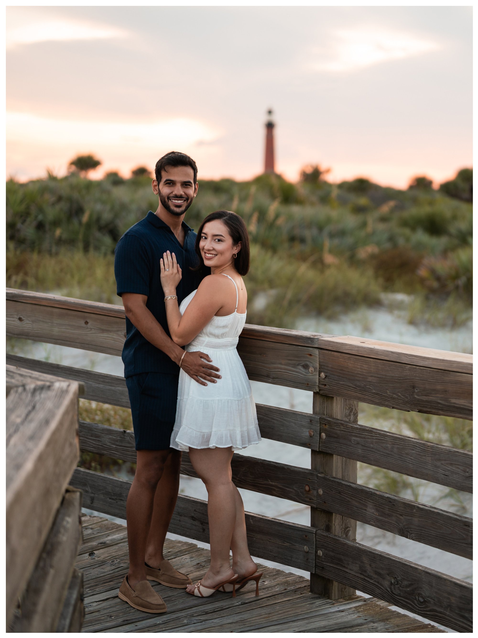 Dierick and Paulina embracing on the boardwalk with the lighthouse behind the dunes.