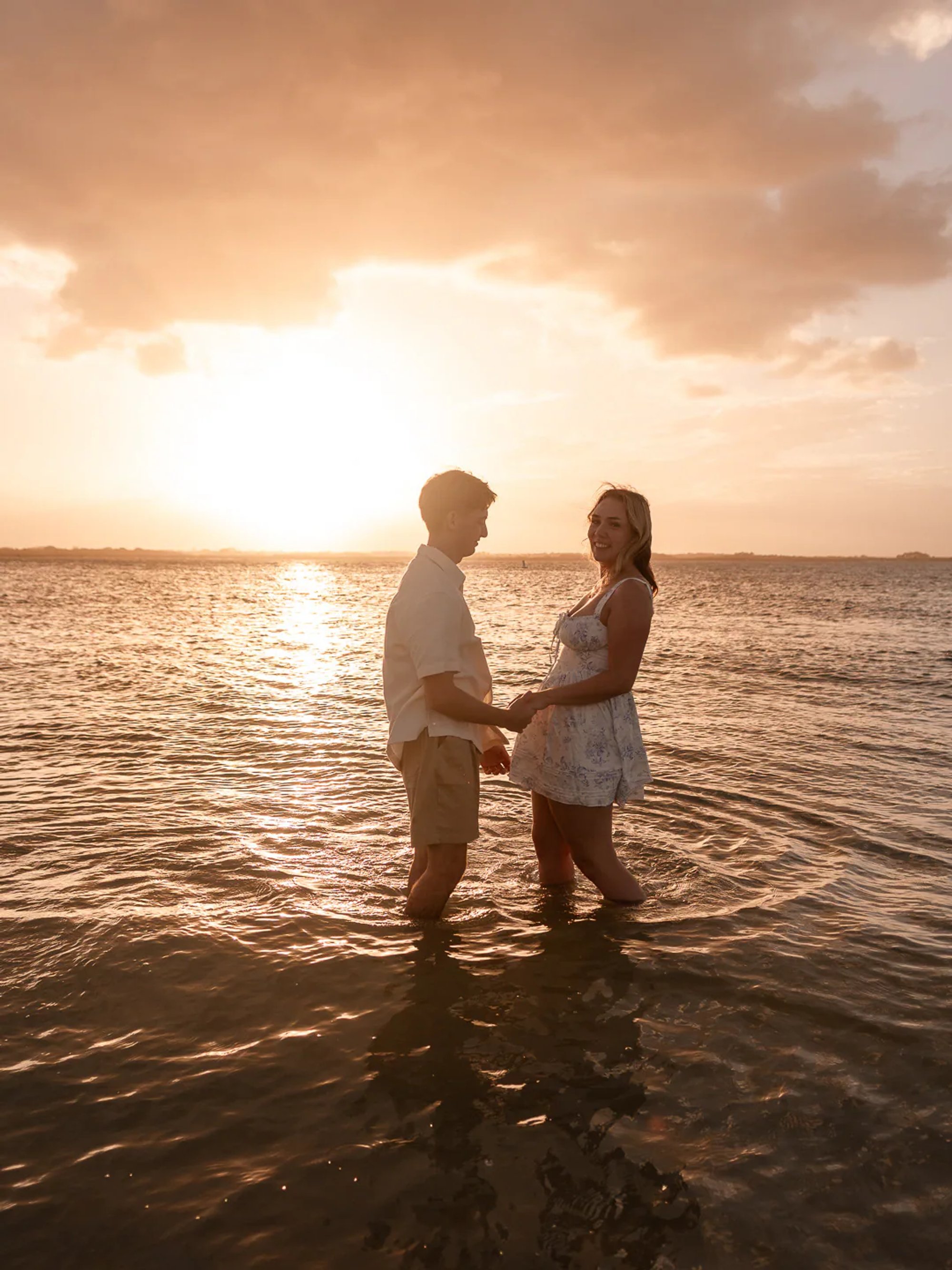 Couple facing each other in shallow water beneath a golden sunset.