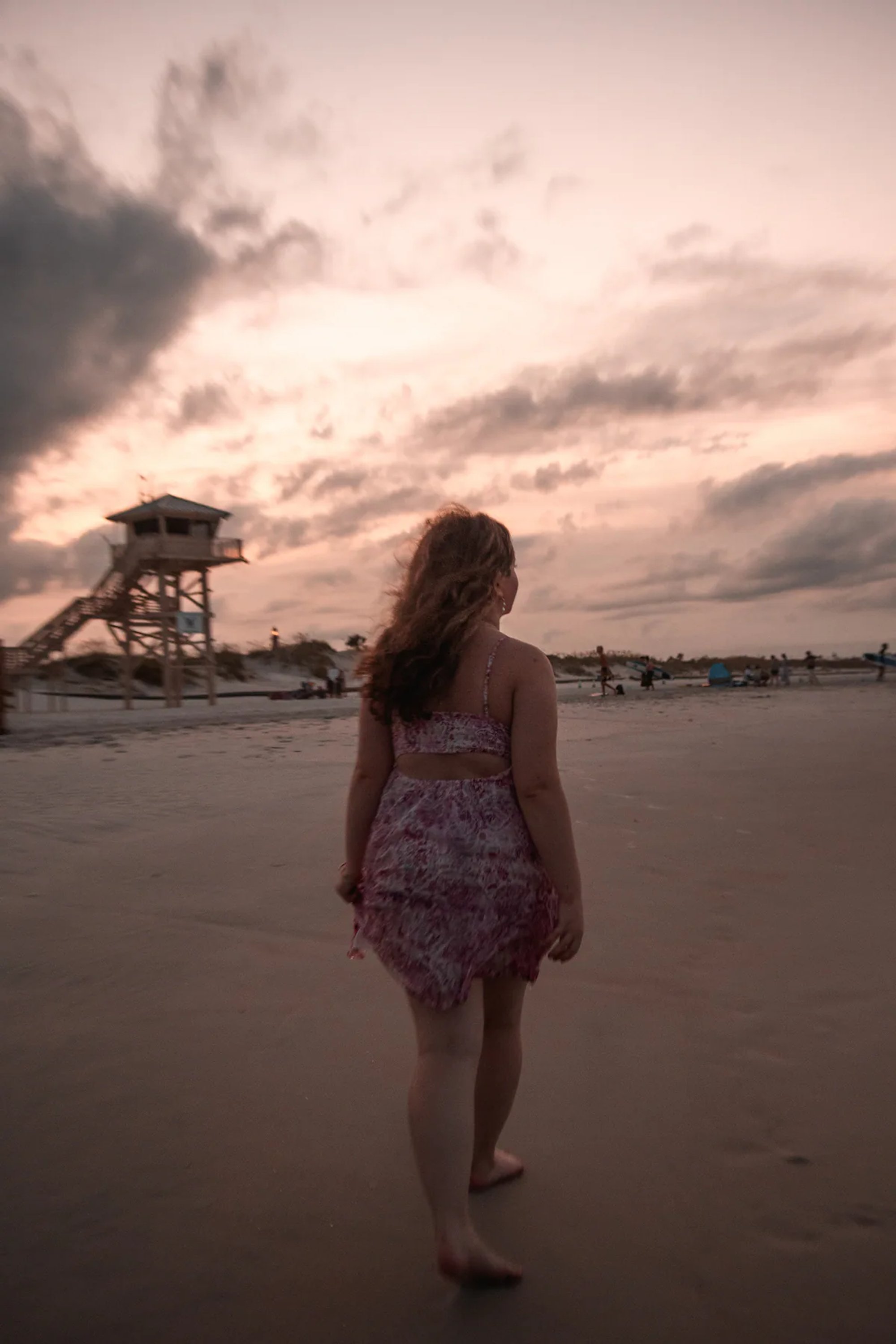 Woman in a pink dress walking on the beach beneath a dramatic sunset sky.