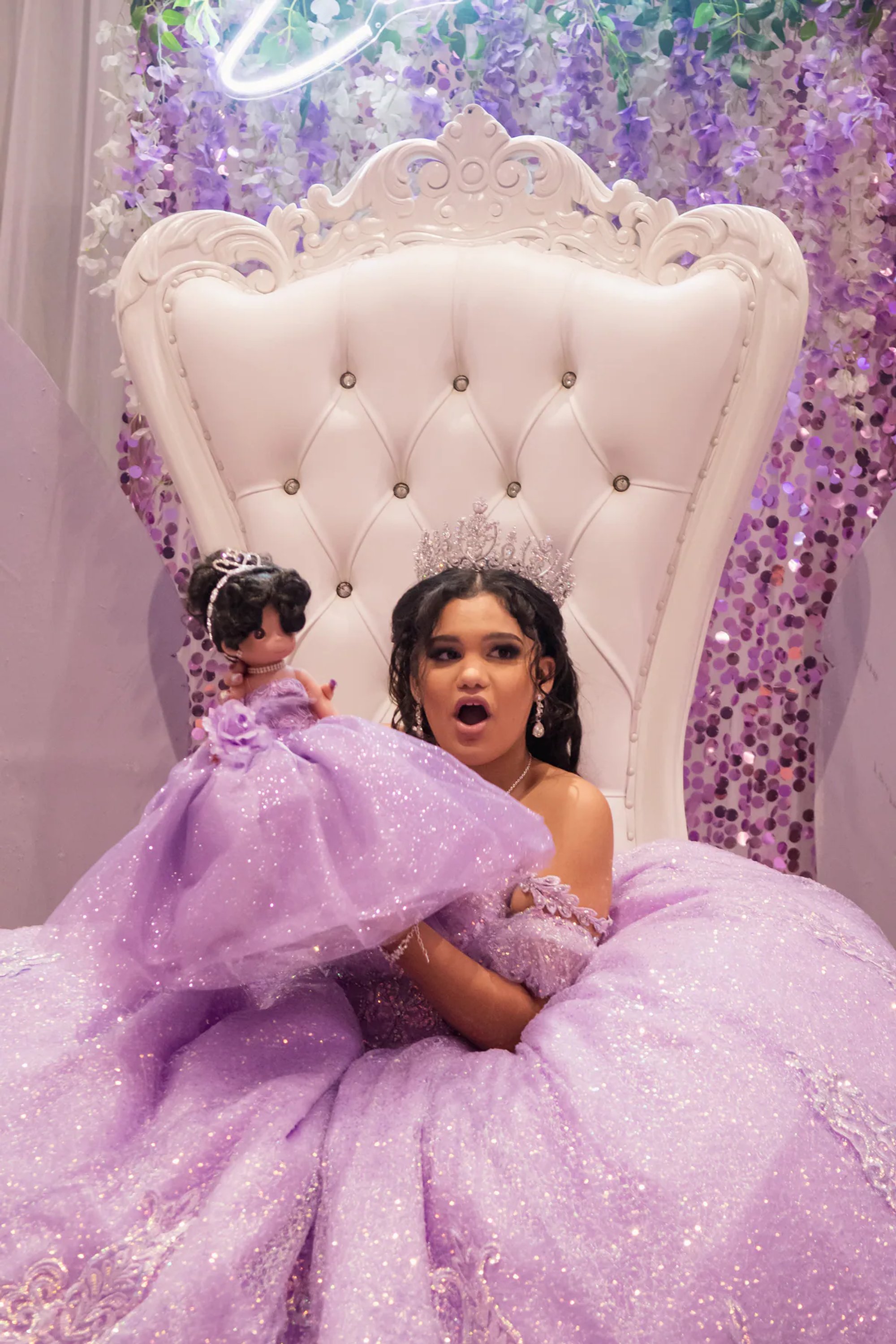 Quinceañera seated on a white throne holding a doll in a matching purple gown.