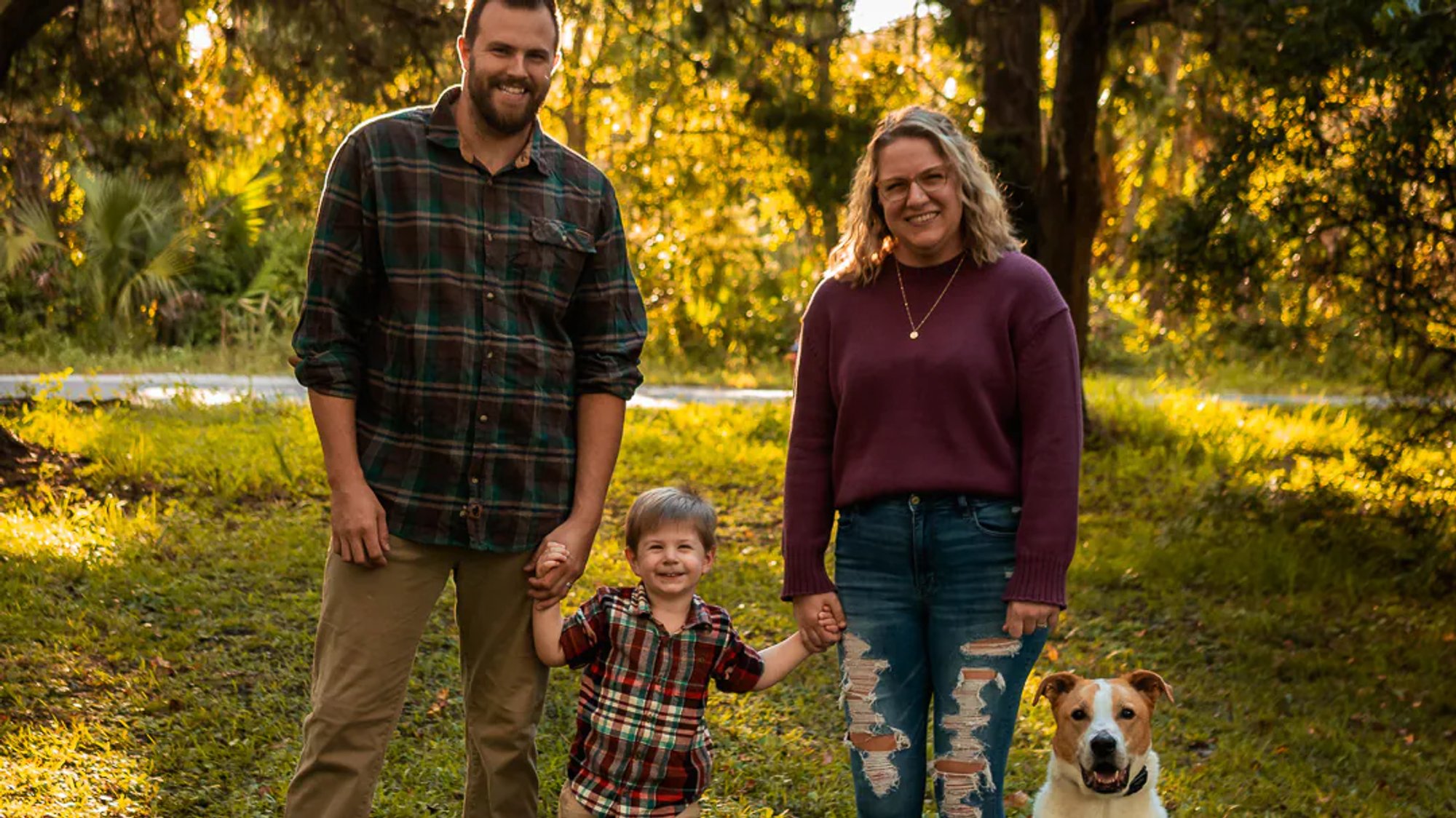 Parents standing with their young child and dog in warm wooded light.