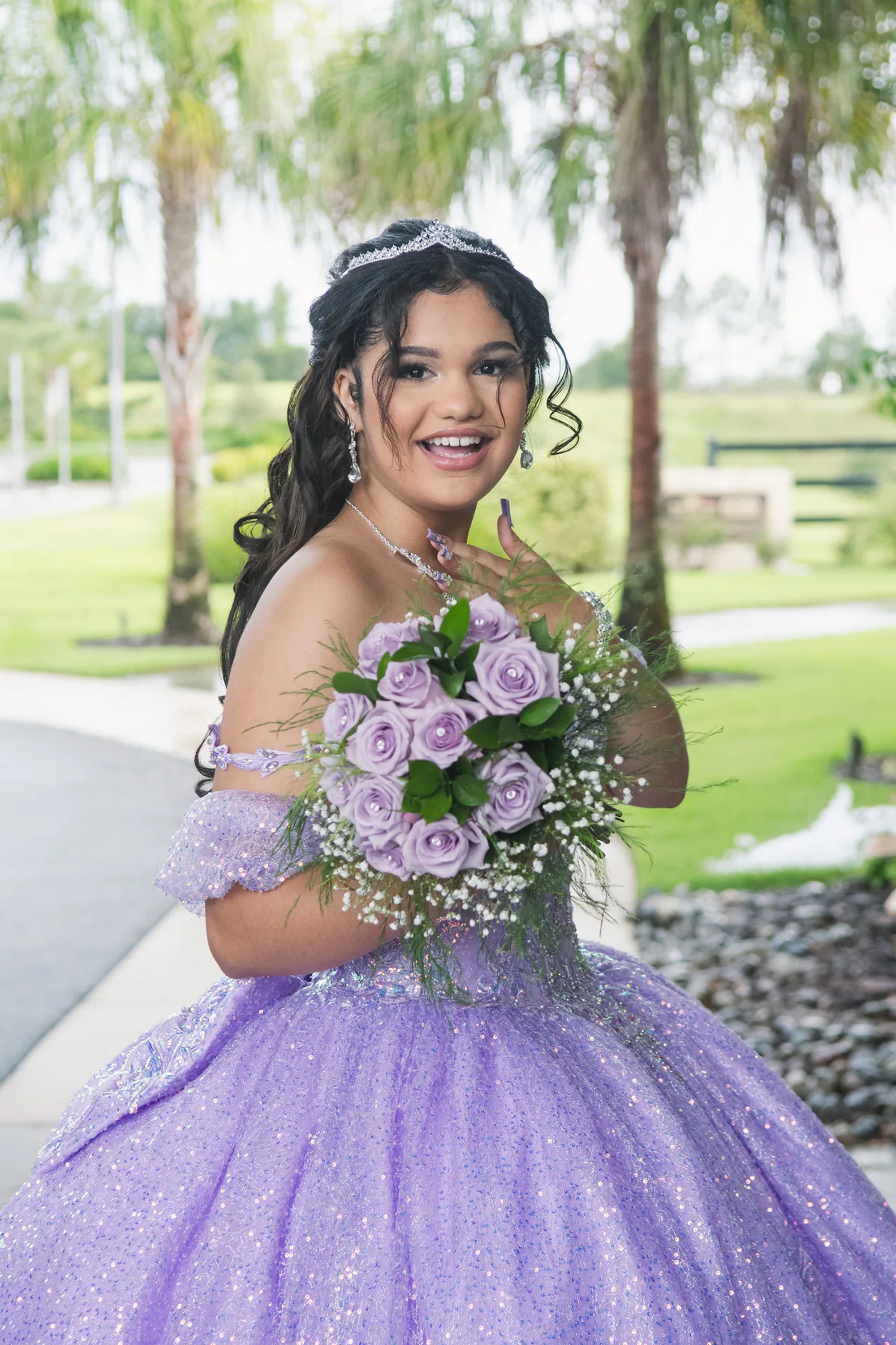 Smiling quinceañera holding her purple bouquet outdoors.