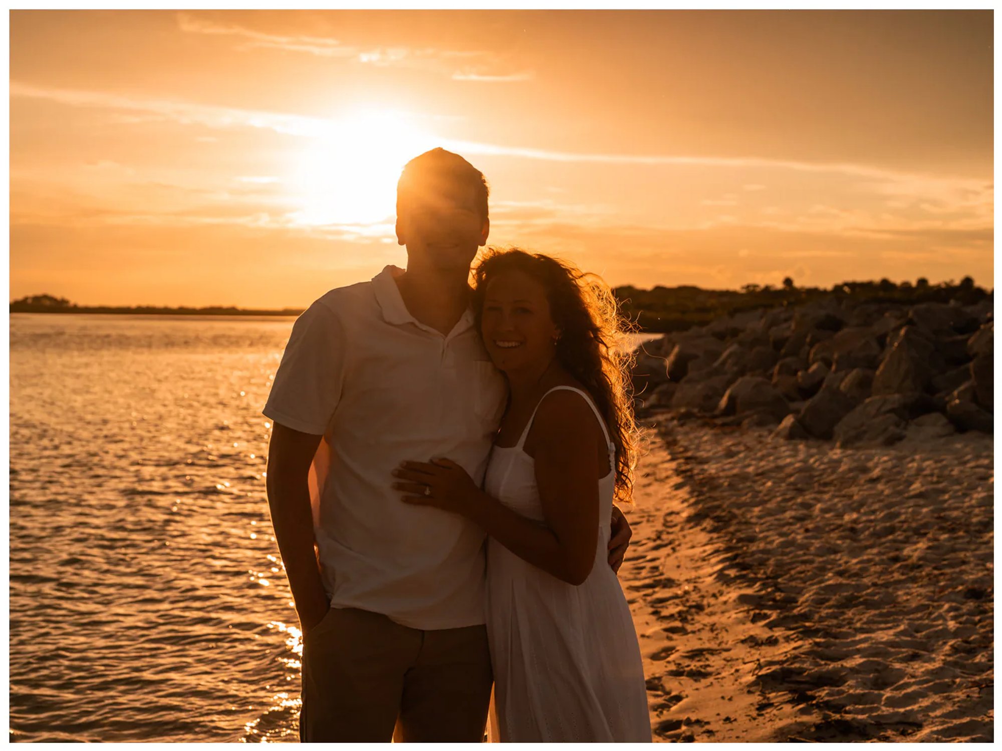 Backlit couple standing close together beside the water.