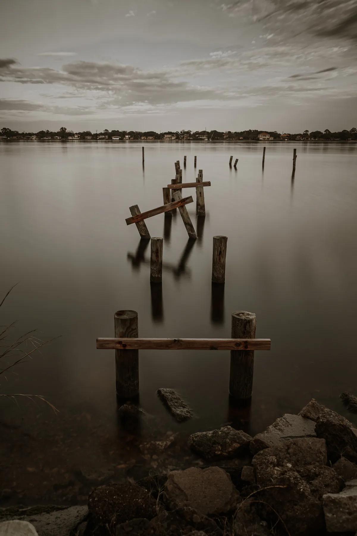 Broken wooden dock posts extending into calm water beneath an overcast sky.