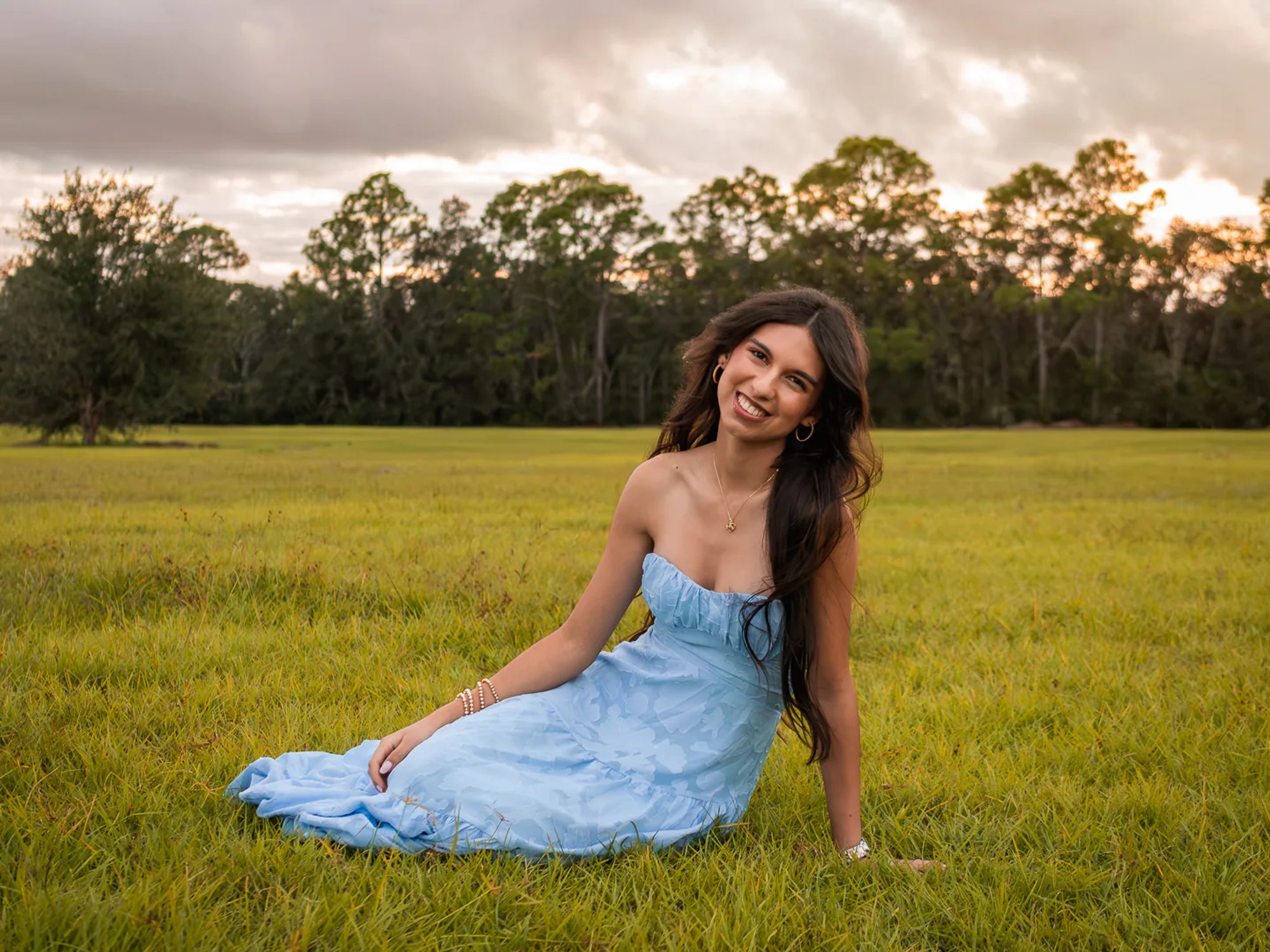 Young woman smiling while seated in a pale blue dress on the grass.