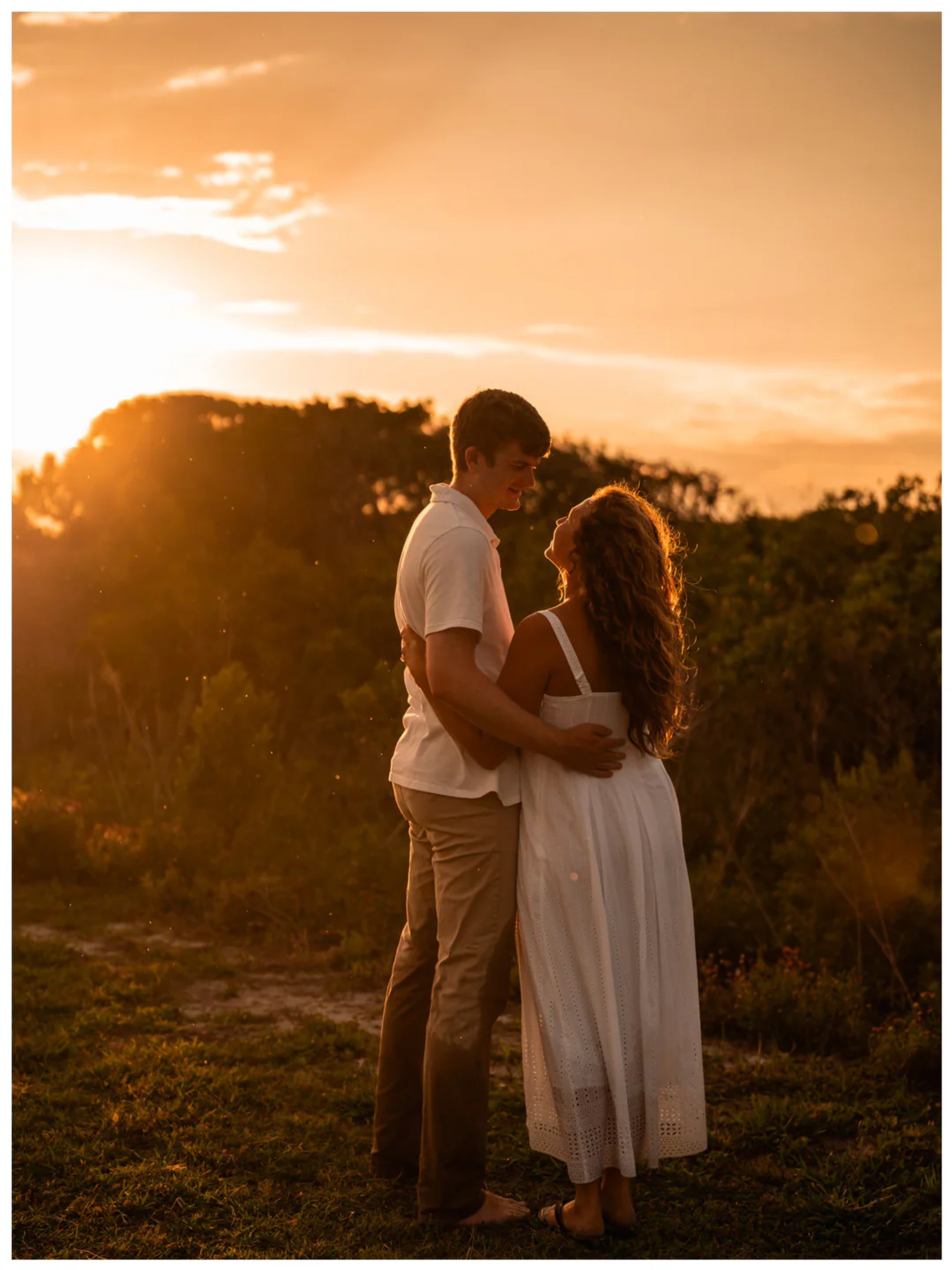 Couple embracing near coastal plants with a lighthouse in the distance.