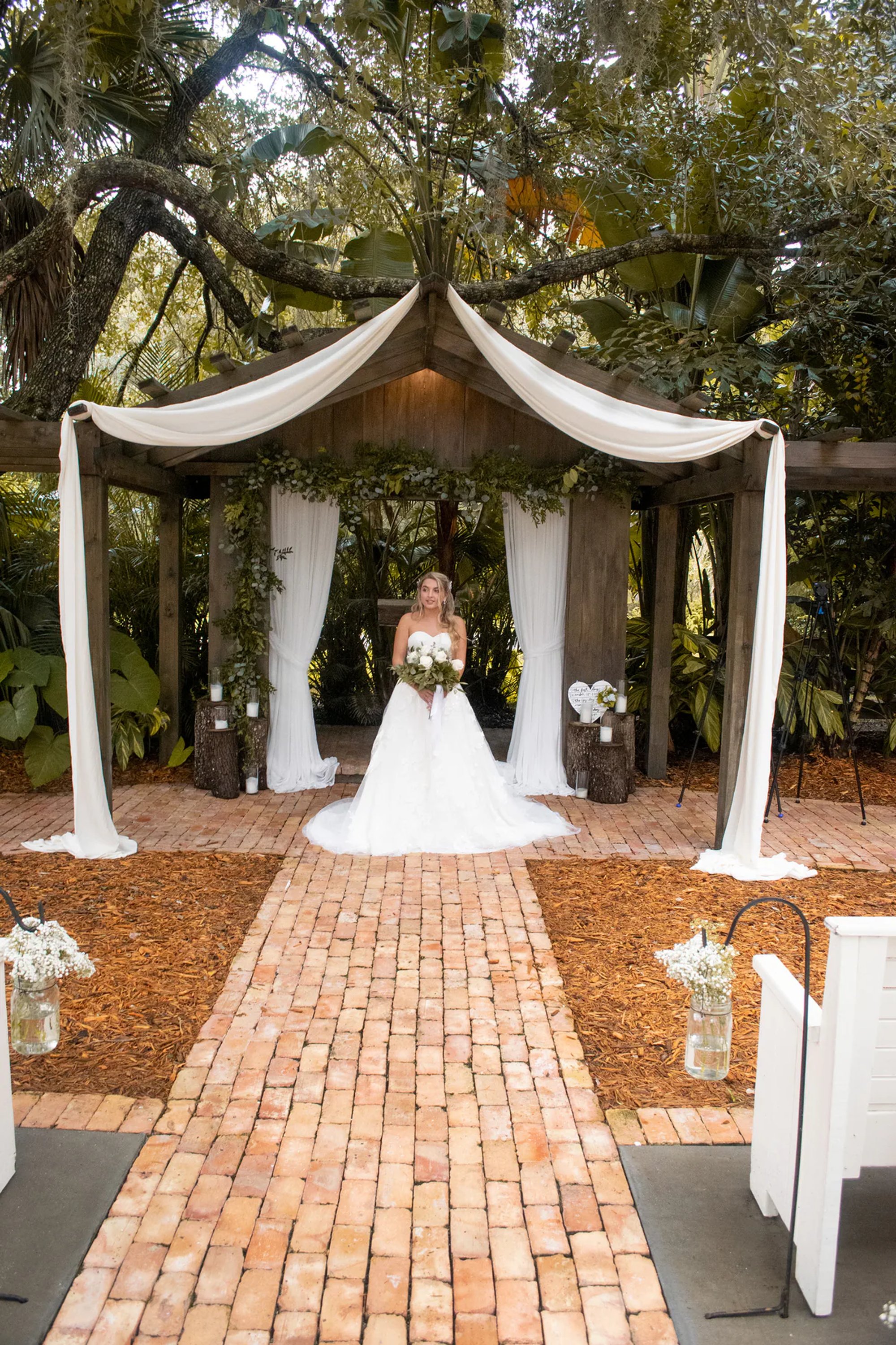 Bride standing beneath a draped outdoor wedding arch at the end of a brick aisle.