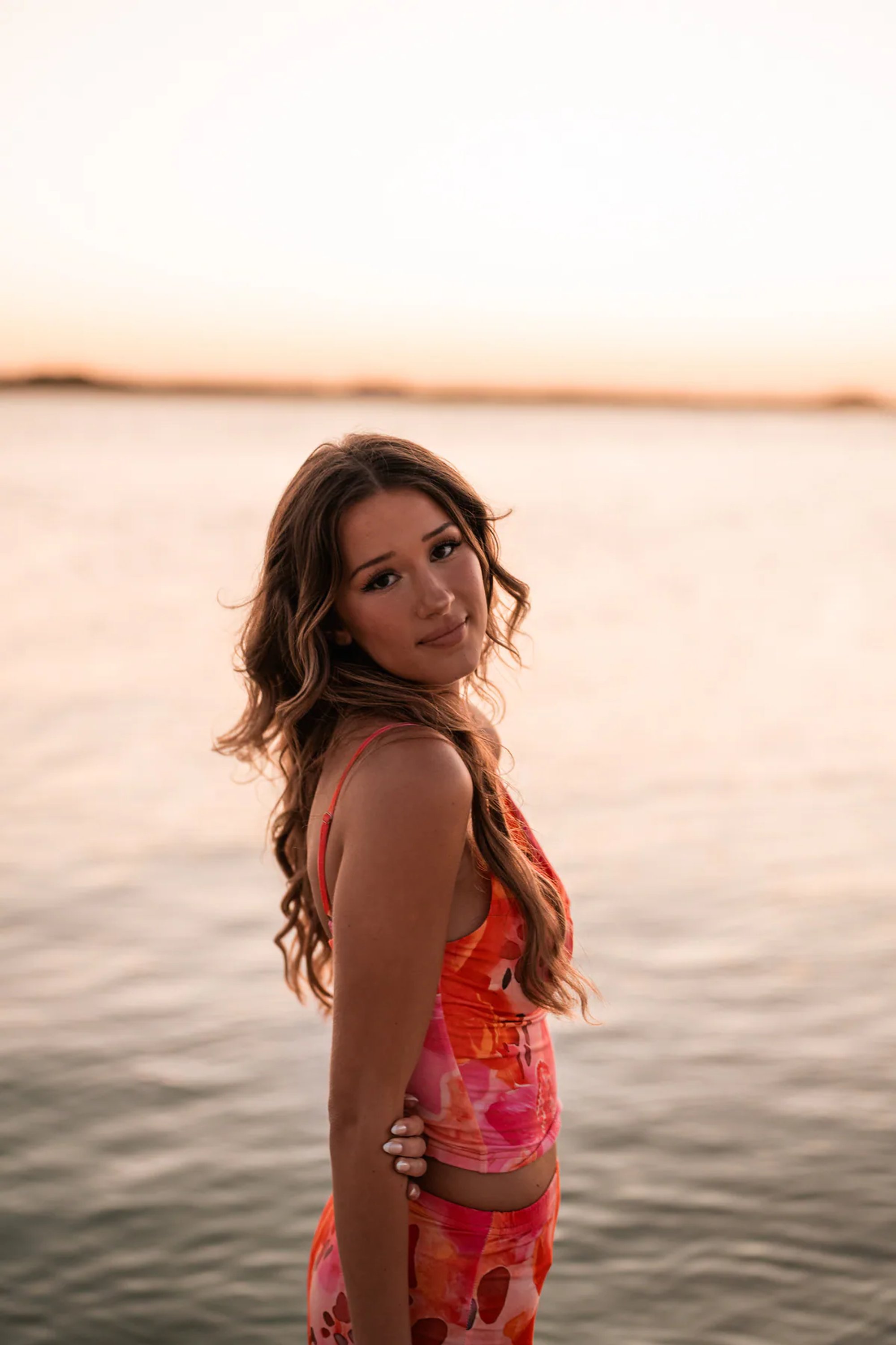 Young woman in a coral patterned dress standing beside calm water.