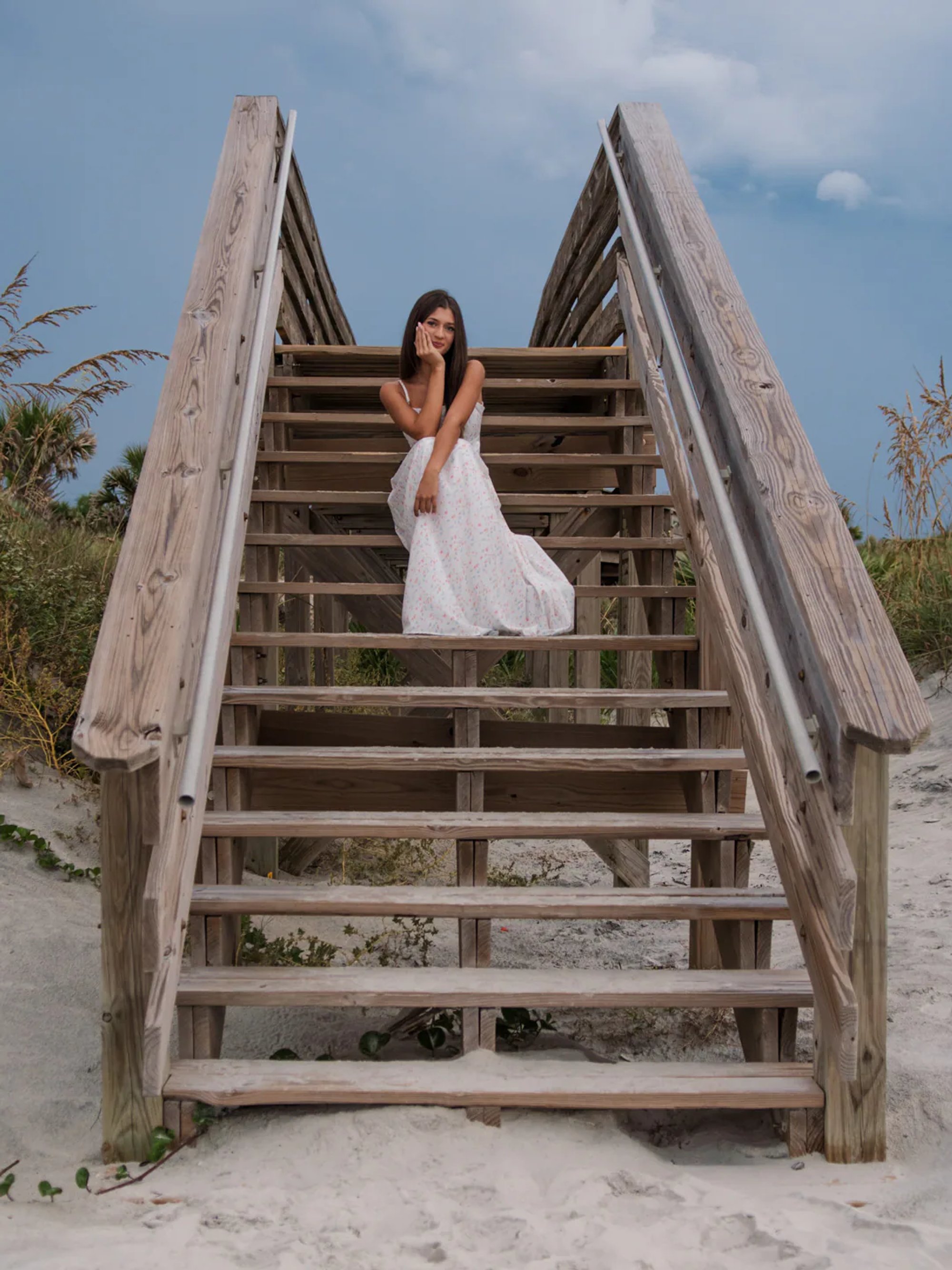 Senior photo session in Ponce Inlet dunes, Mia sitting on wooden beach stairs in a white floral maxi dress.