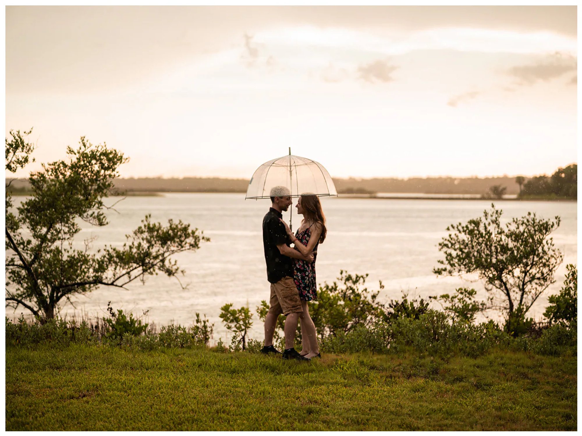 Wide portrait of a couple embracing beneath an umbrella on the grassy waterfront at Washington Oaks Gardens.