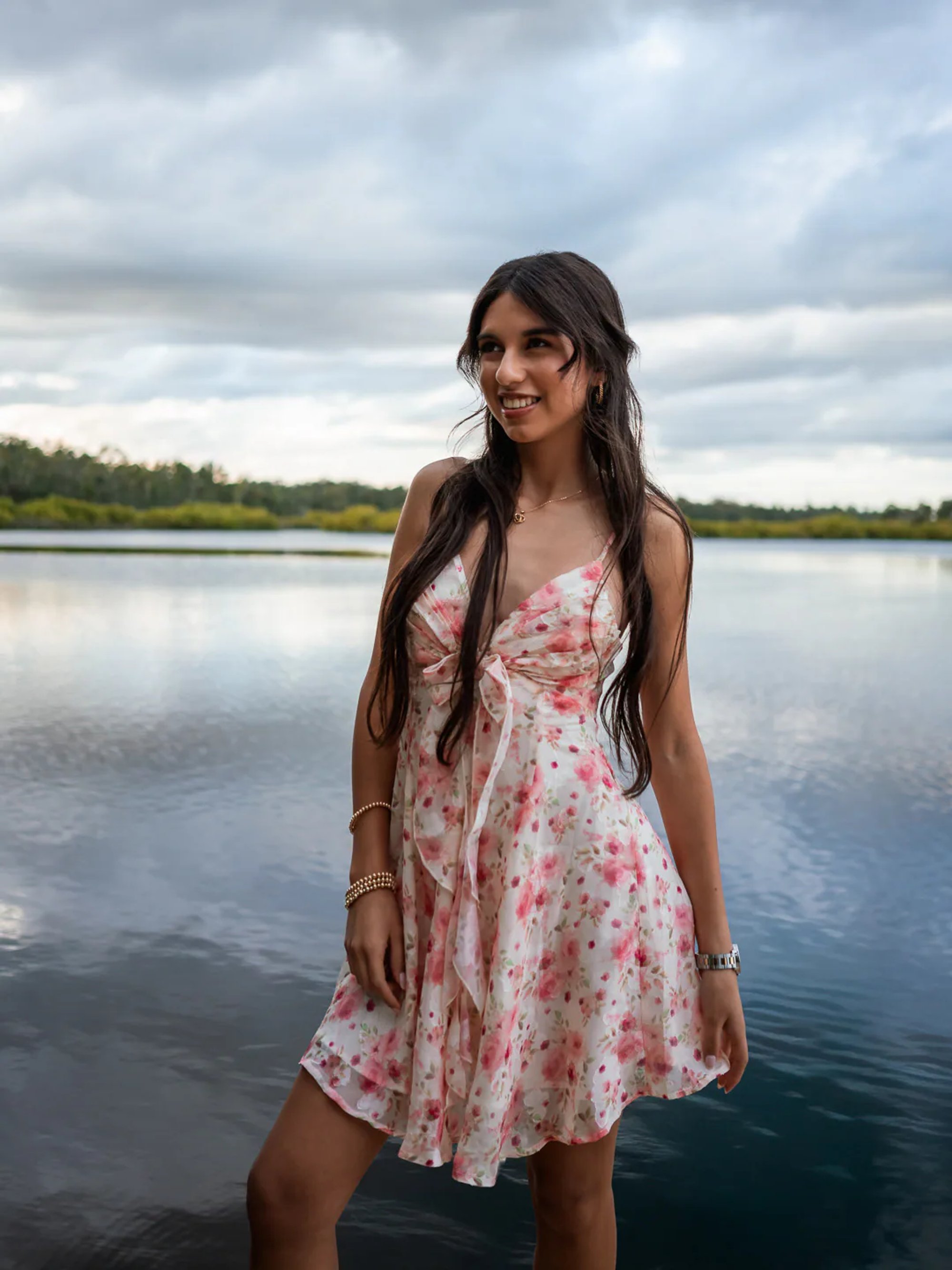 Young woman in a floral dress standing beside a calm lake.