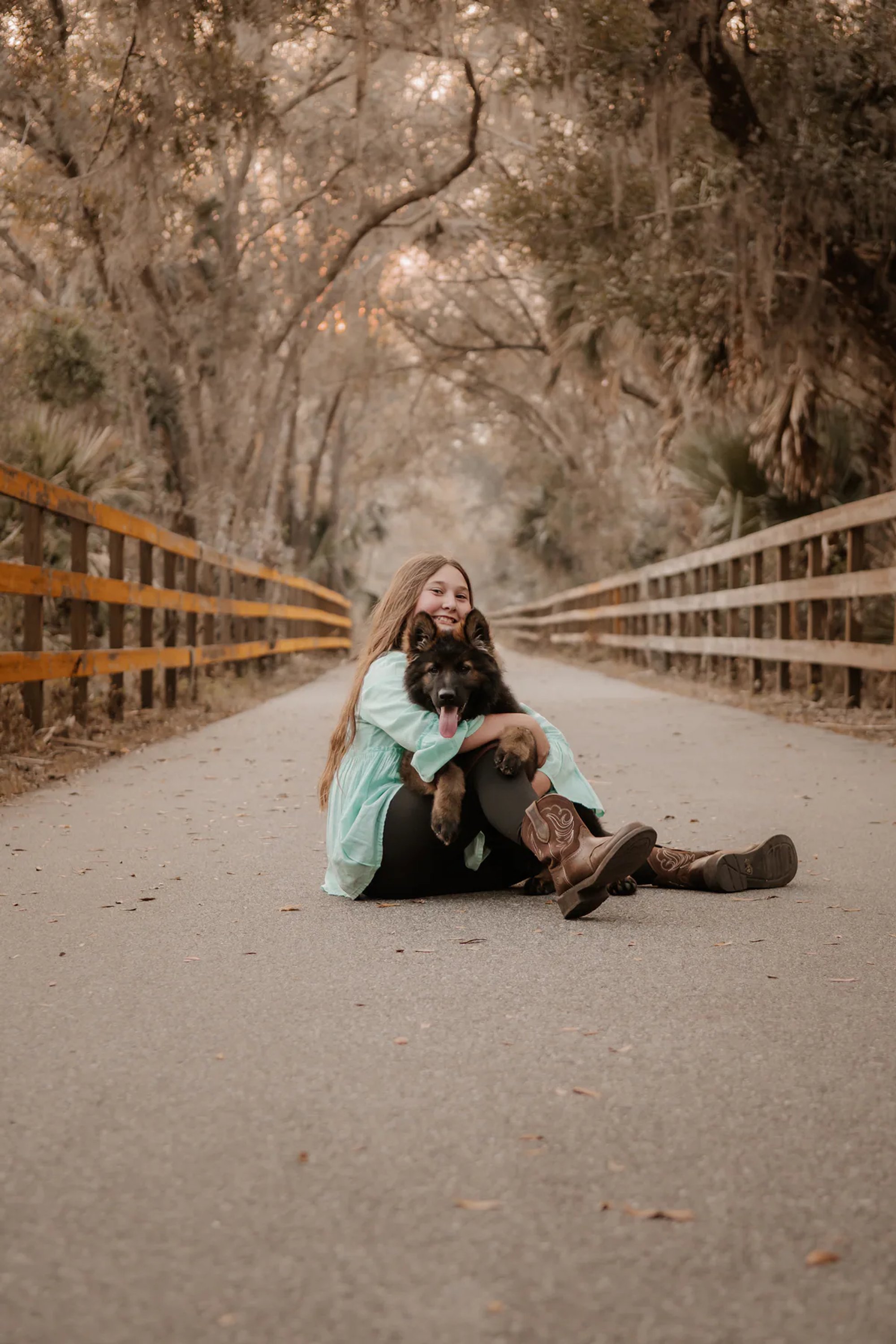 Young woman seated with her black dog beneath arching oak trees.