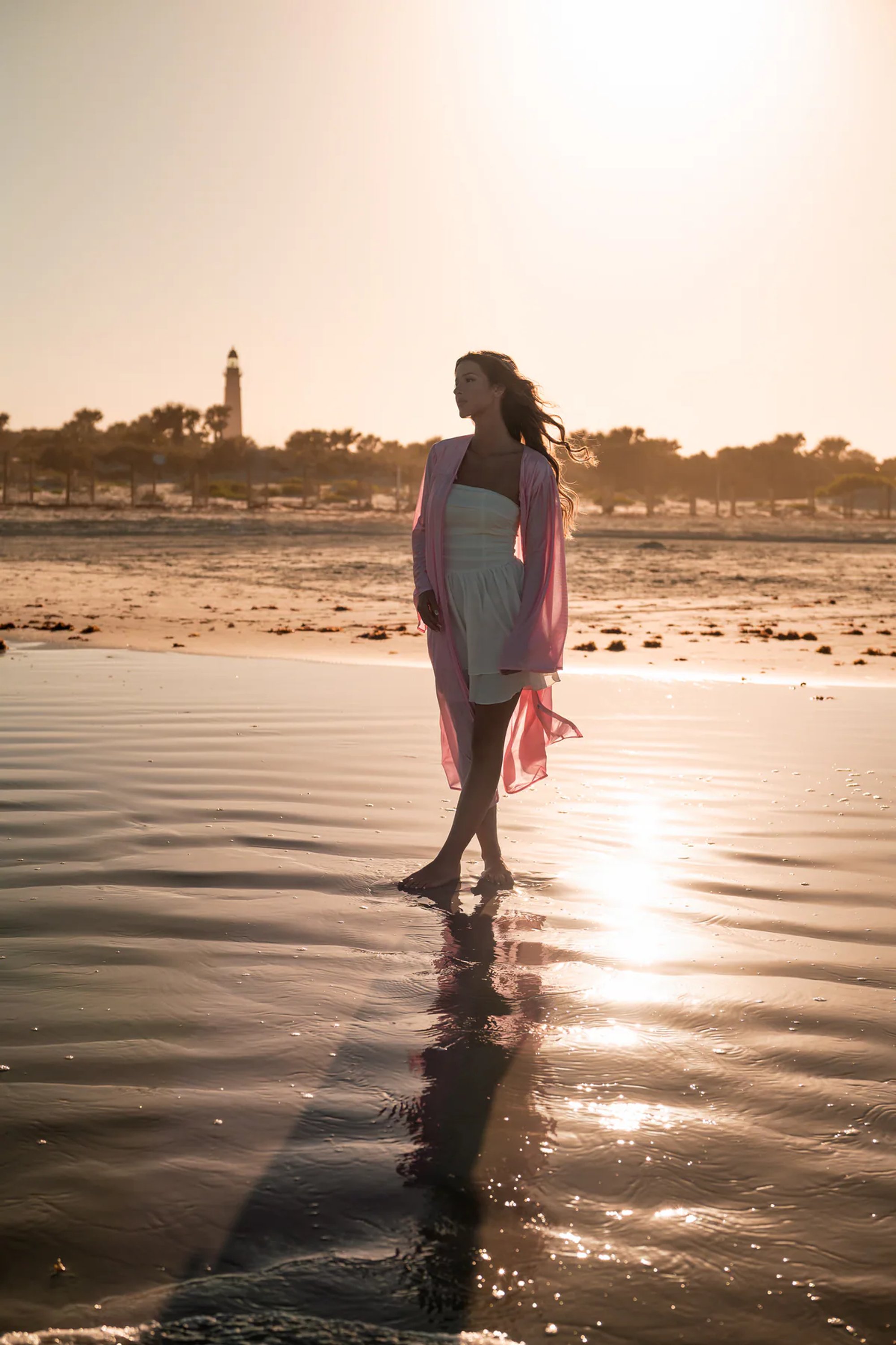 Backlit young woman in a pink wrap standing on reflective wet sand.