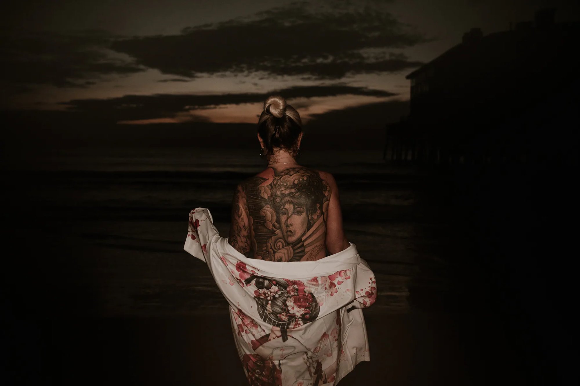 Tattooed woman holding a floral wrap with her back to the camera at a dark shoreline.