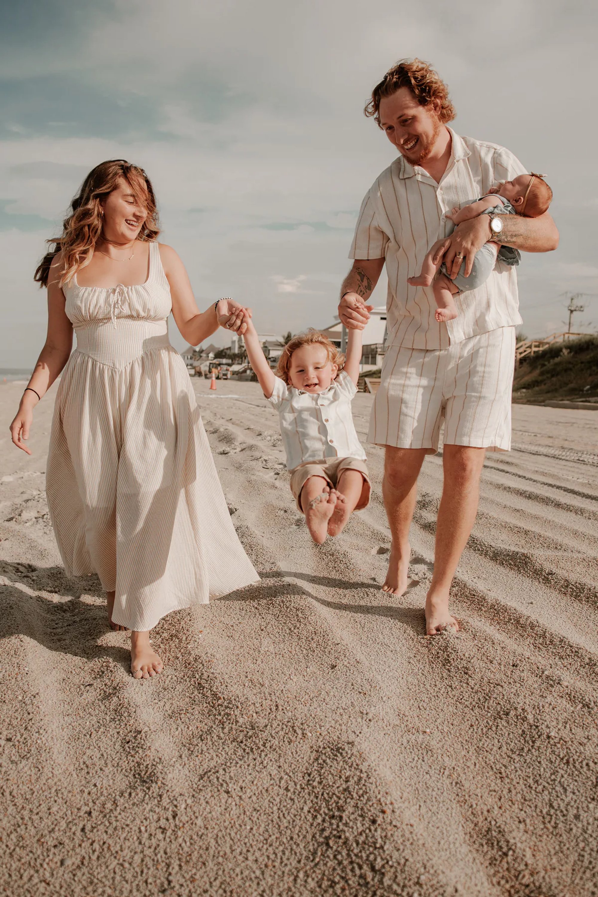 Parents walking along the sand while holding their toddler's hands.
