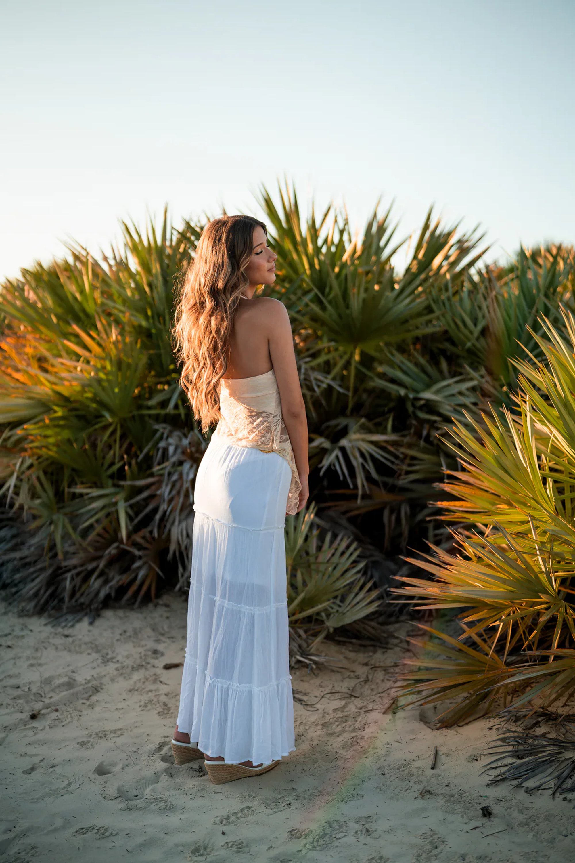 Young woman in a long white dress standing beside sunlit palmettos.