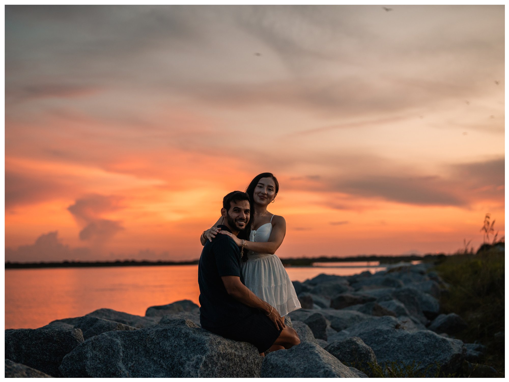 Dierick and Paulina sitting together on the rocks beneath a vivid orange sunset.