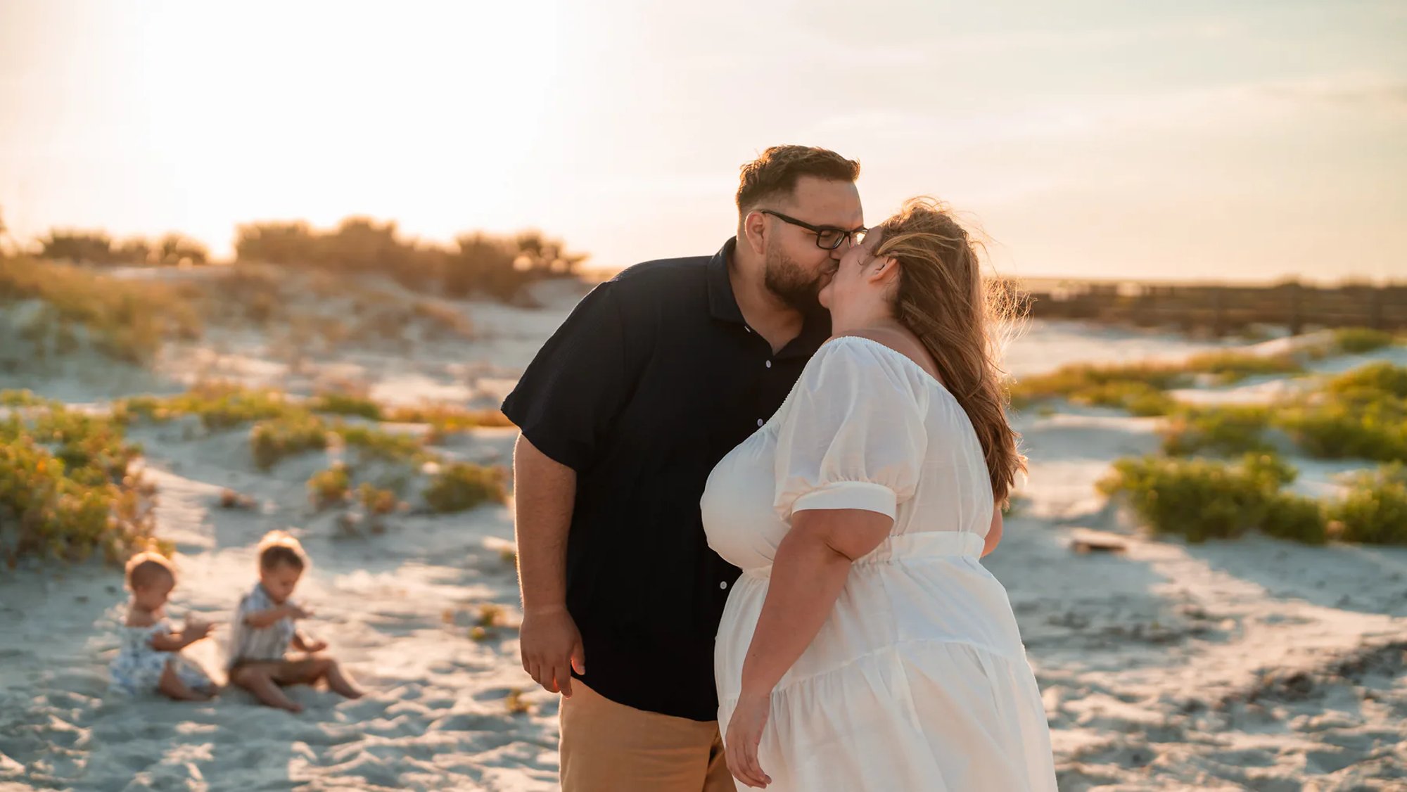 Couple kissing among coastal dunes while two children sit in the background.