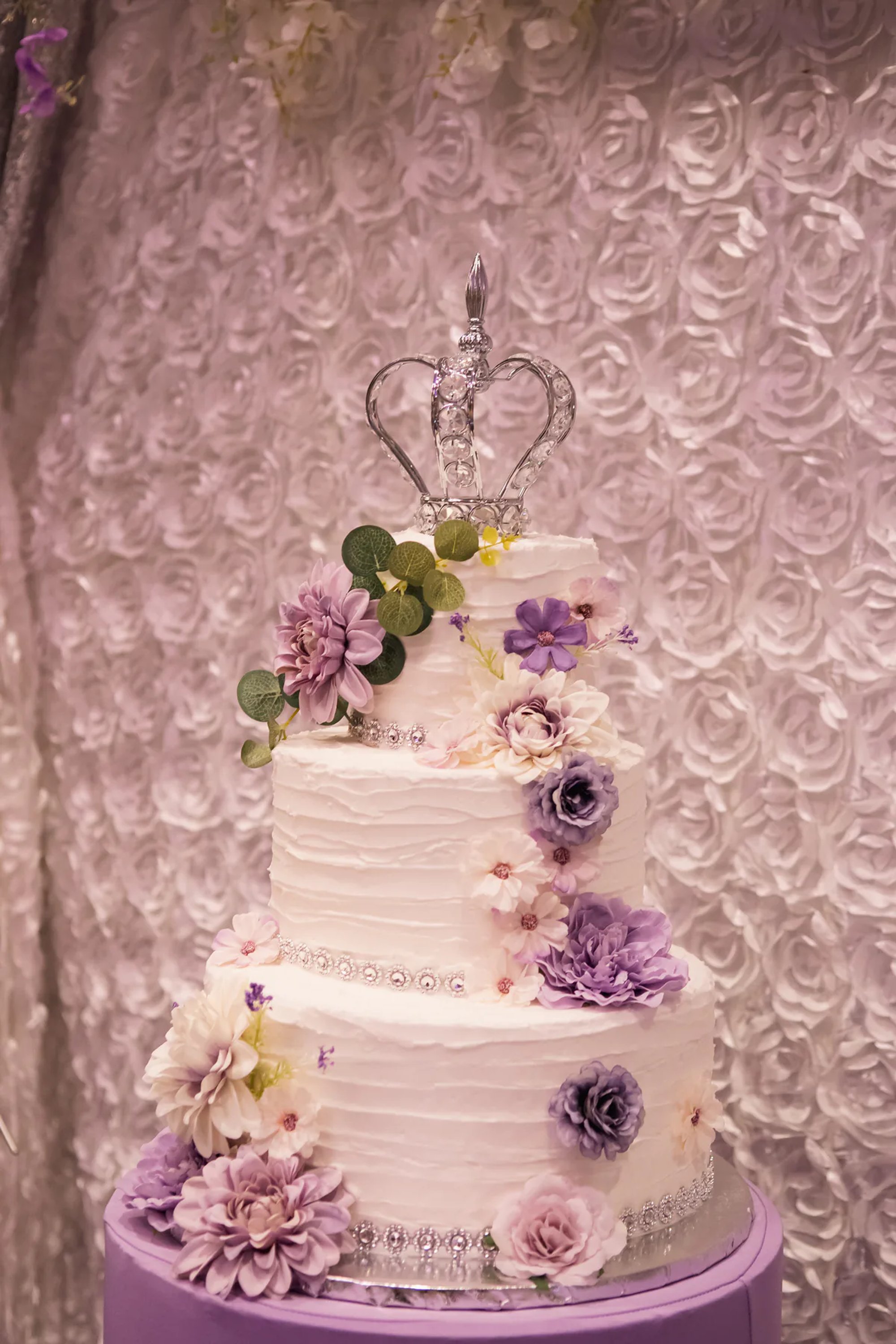 Tiered quinceañera cake decorated with purple flowers and a small crown.
