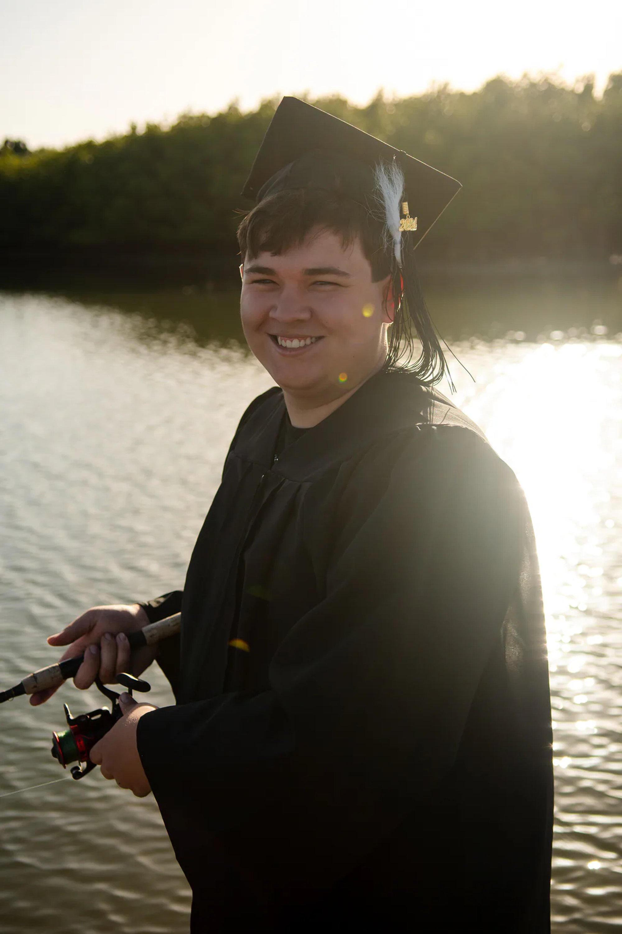 Graduate in a black cap and gown holding a camera beside the water.