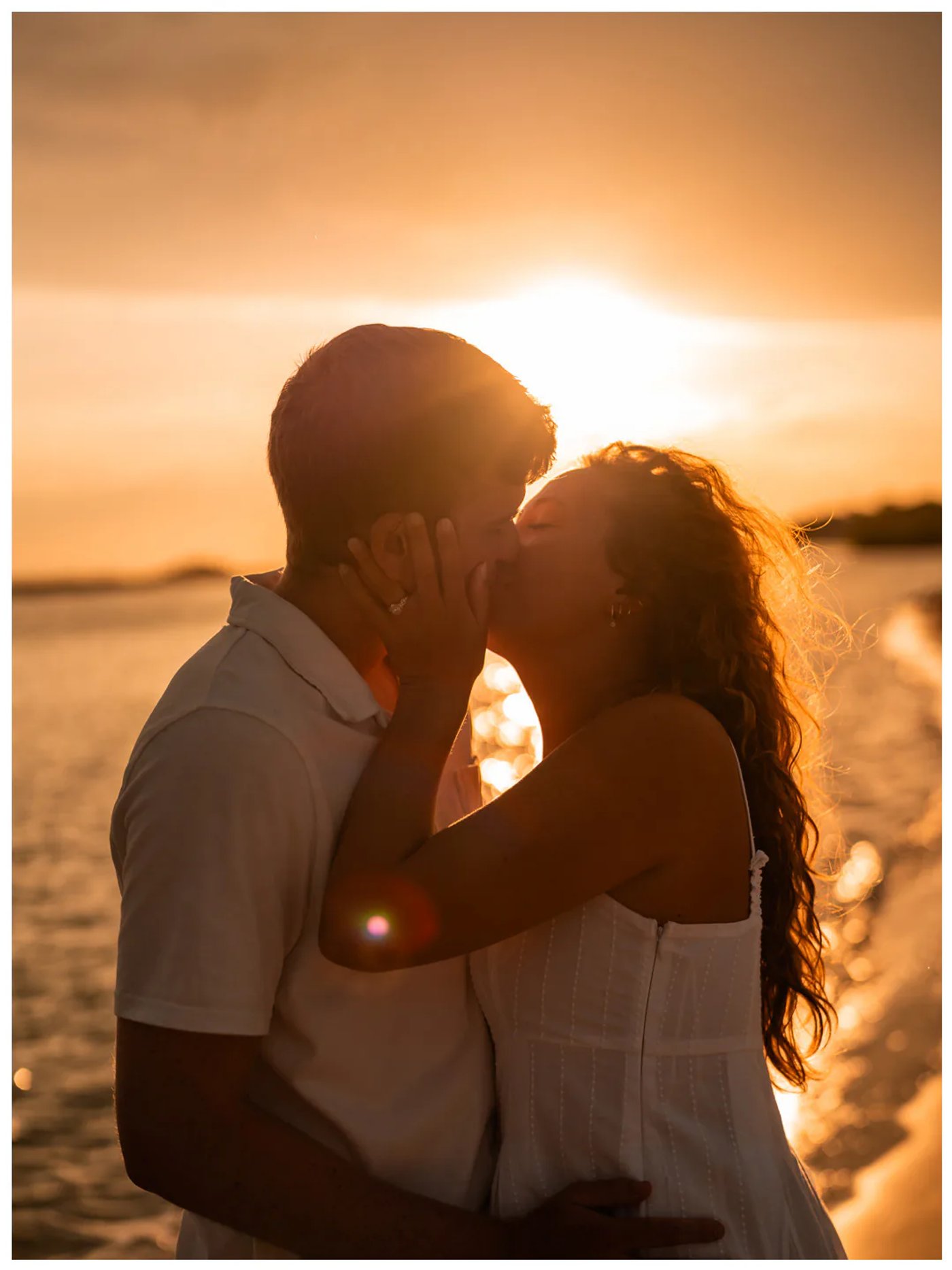 Backlit couple kissing beside the water at sunset.
