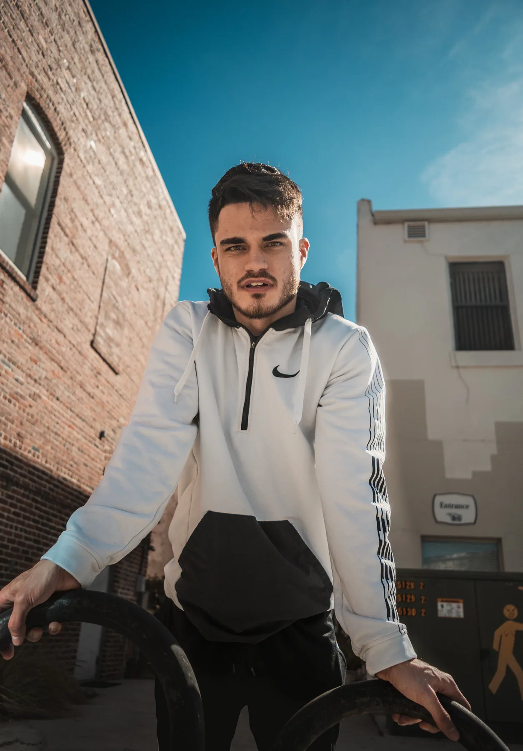 Man in a white hoodie gripping outdoor exercise bars between buildings.