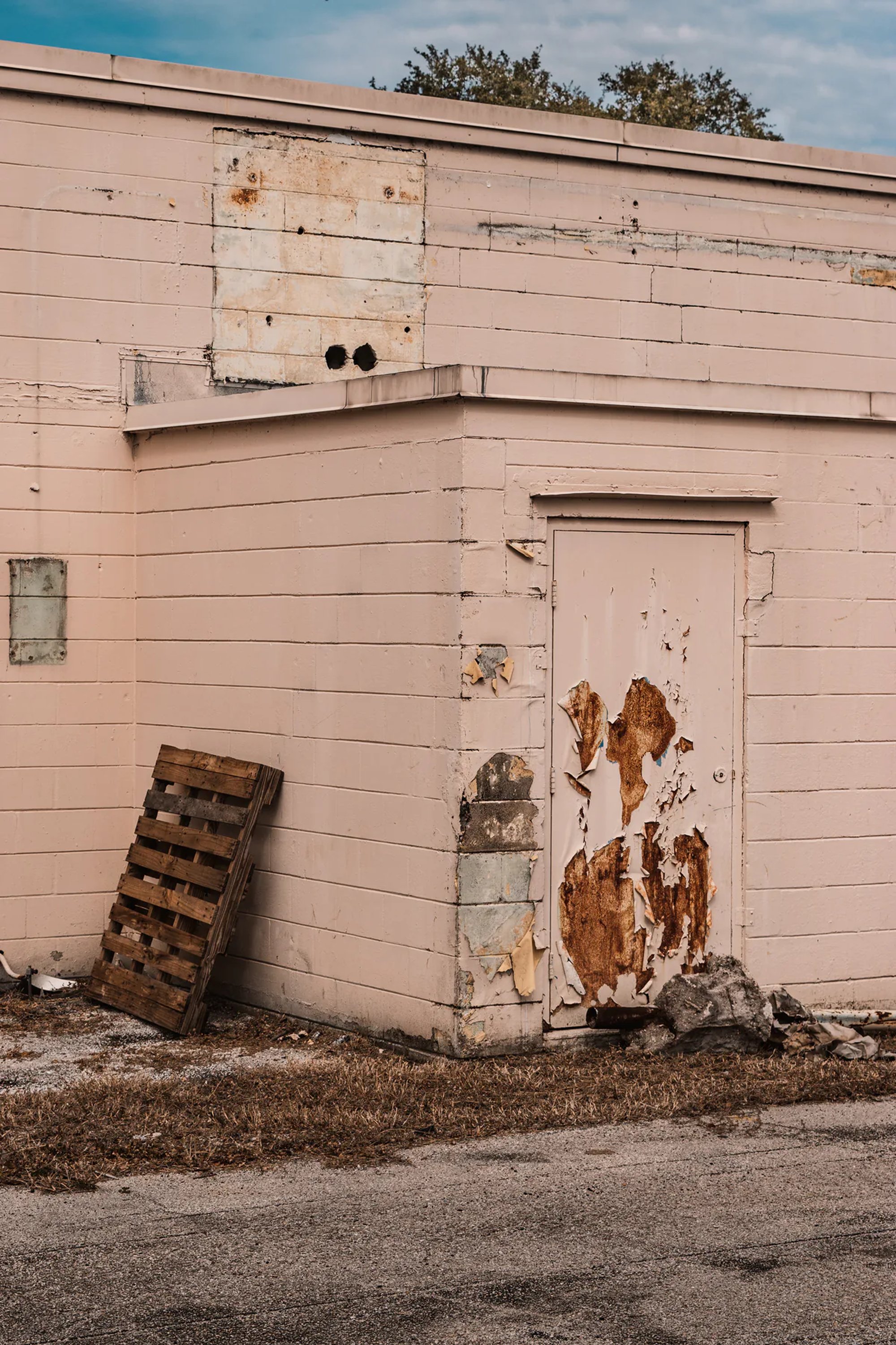 Weathered pale pink building with a worn door and wooden pallet.