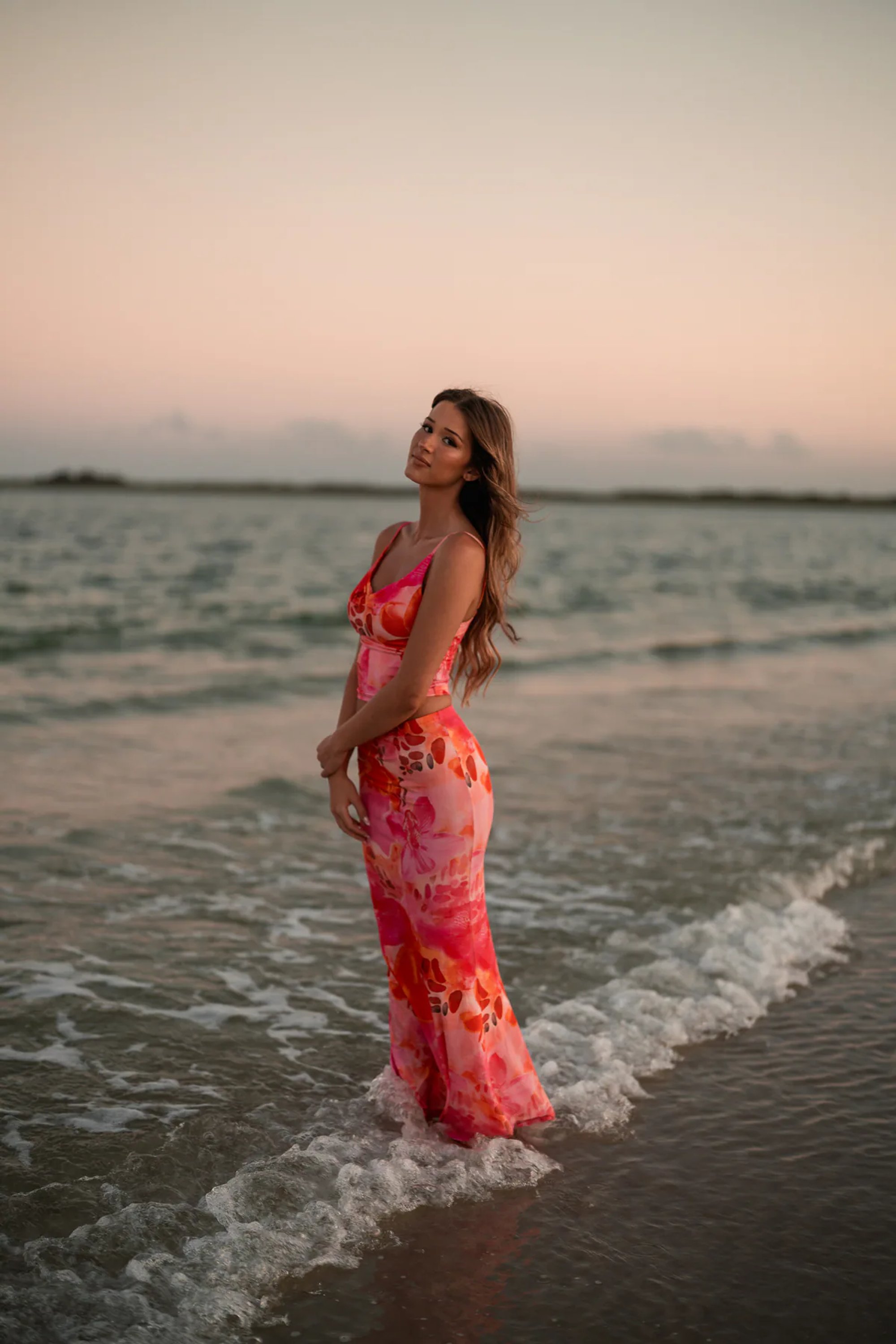 Young woman in a coral patterned dress standing at the edge of the surf.