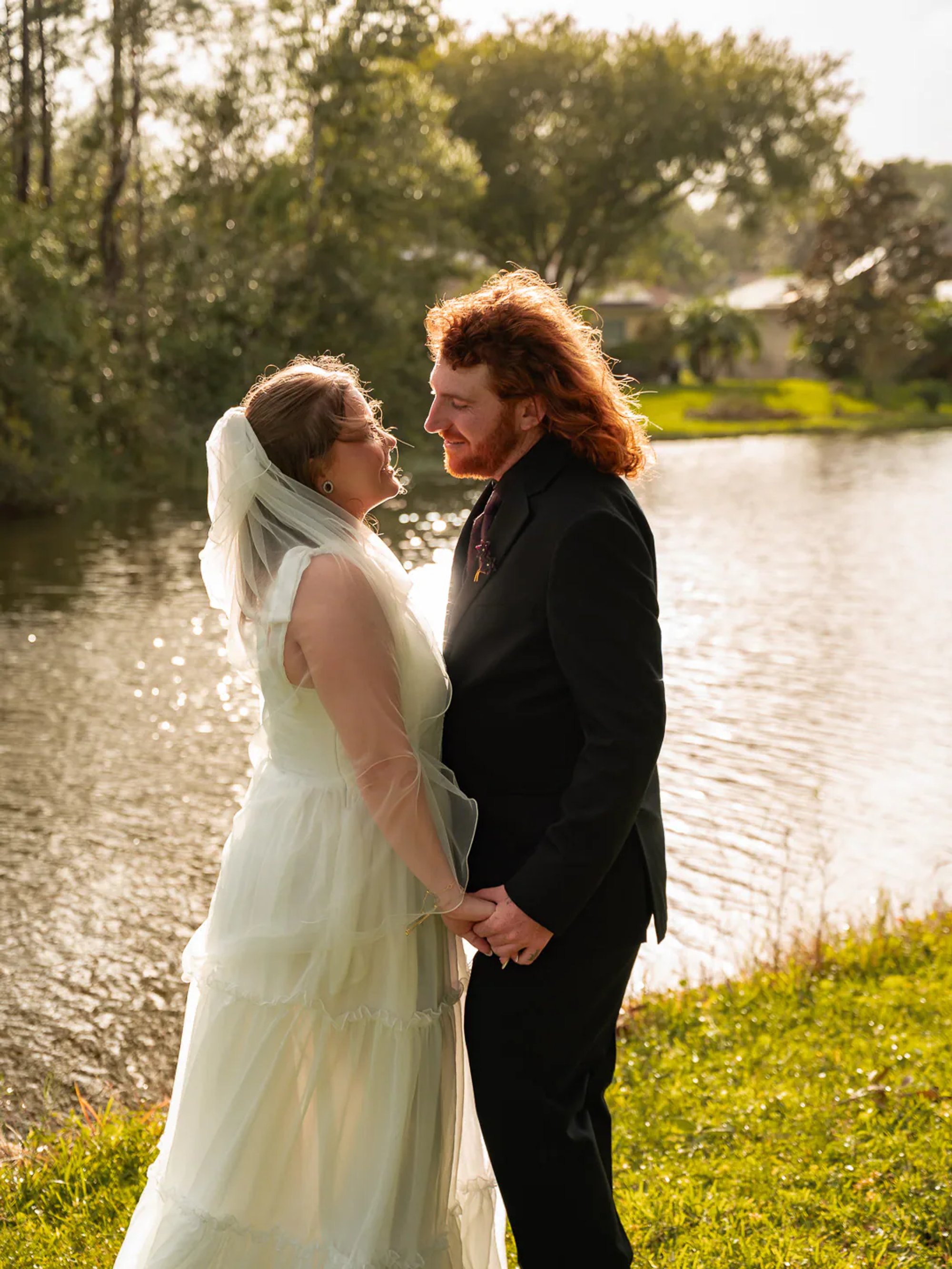 Bride and groom holding hands beside a sunlit pond.
