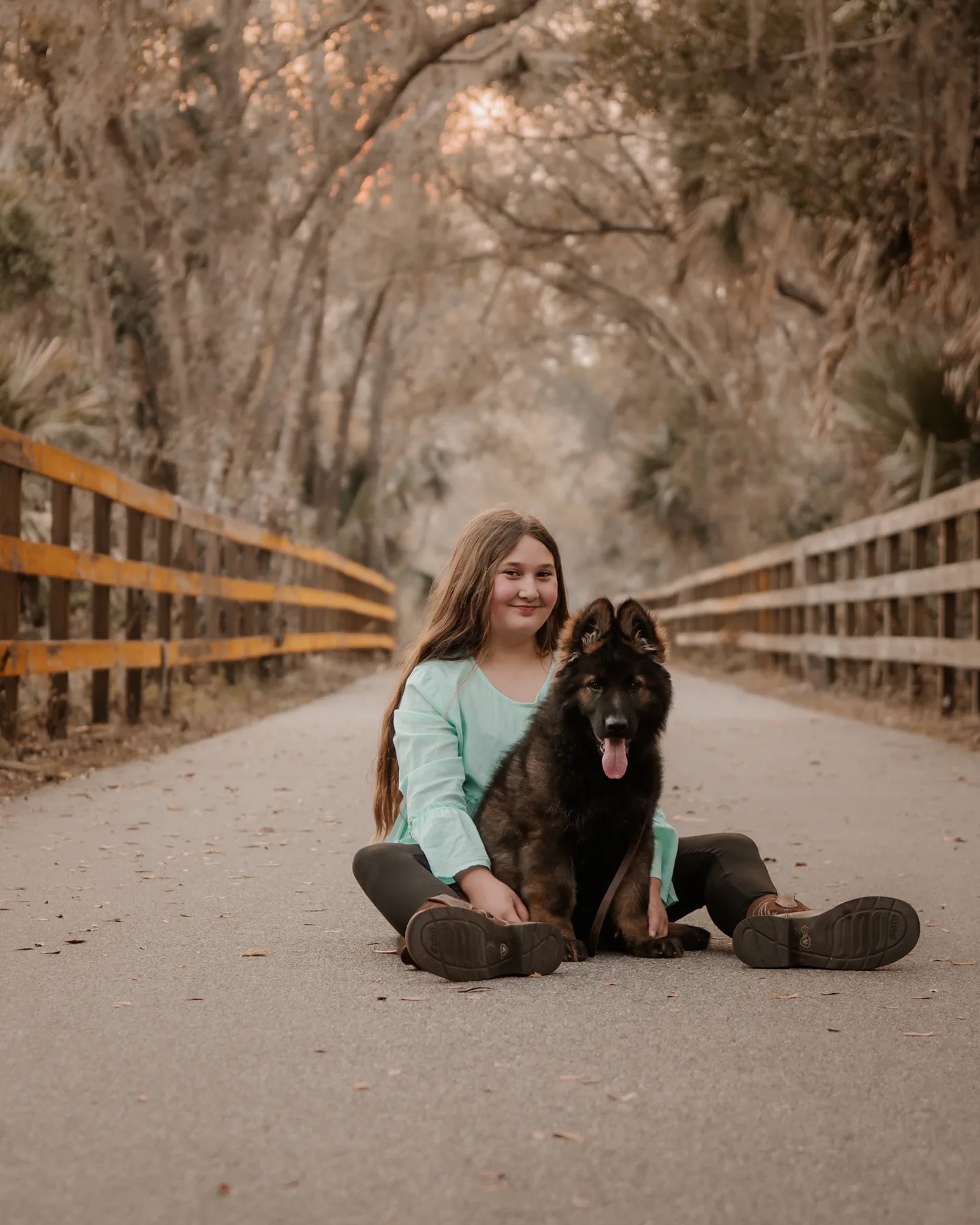 Young woman and her black dog seated together on a tree-lined road.