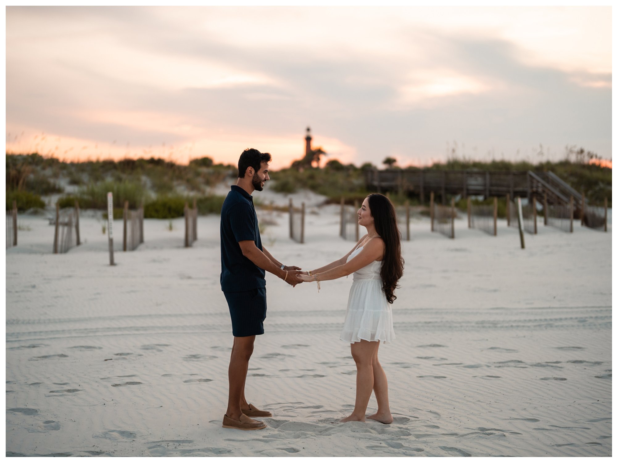 Dierick and Paulina holding hands on the beach beneath a pastel evening sky.