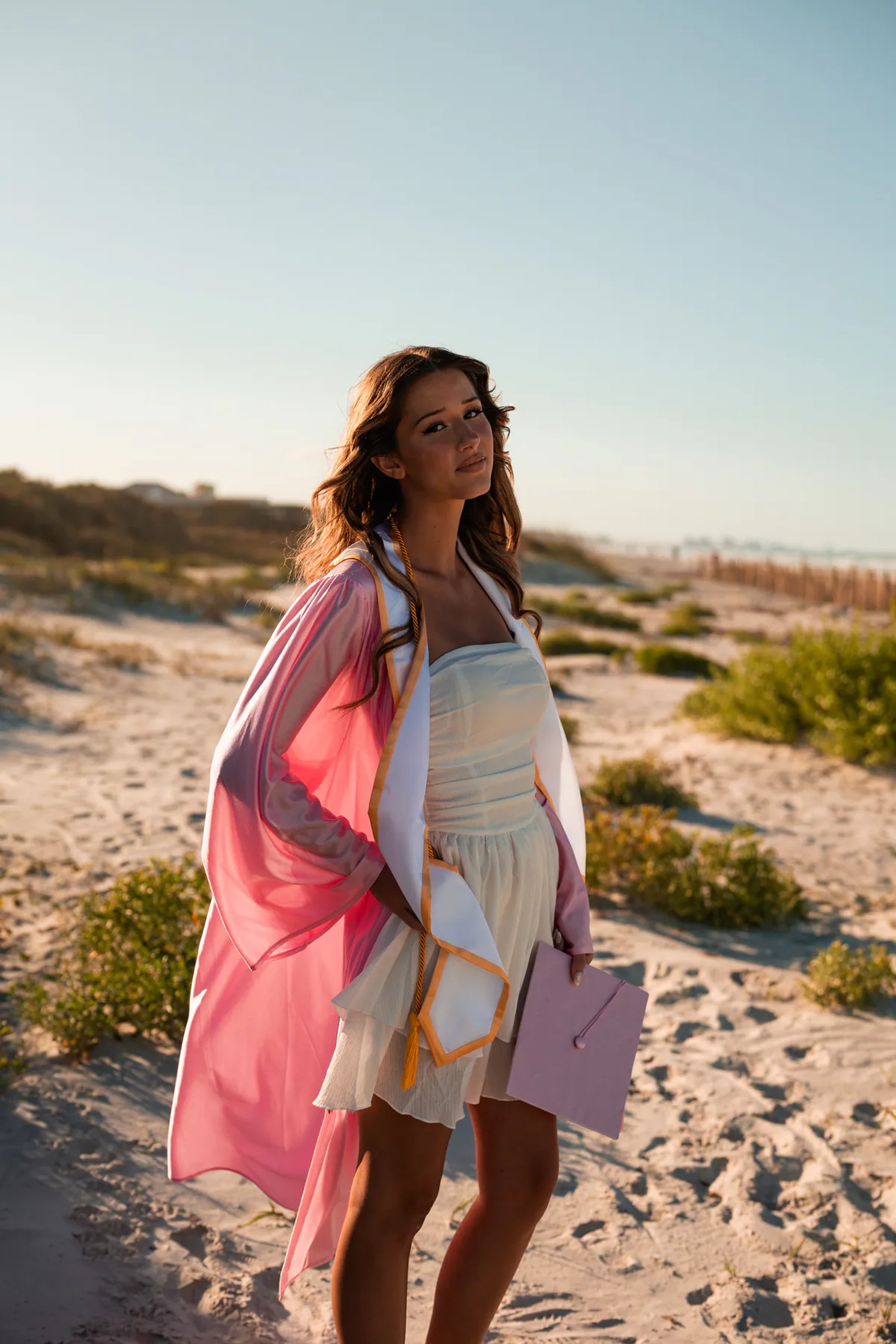 Young woman in a pink wrap and white dress walking near the ocean.
