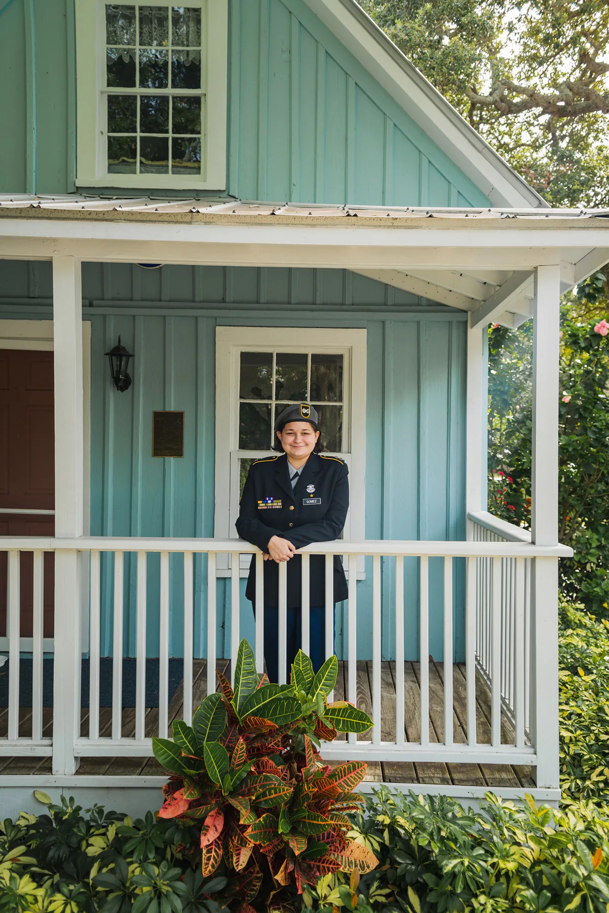 Young person in a dress uniform standing on the porch of a blue cottage.