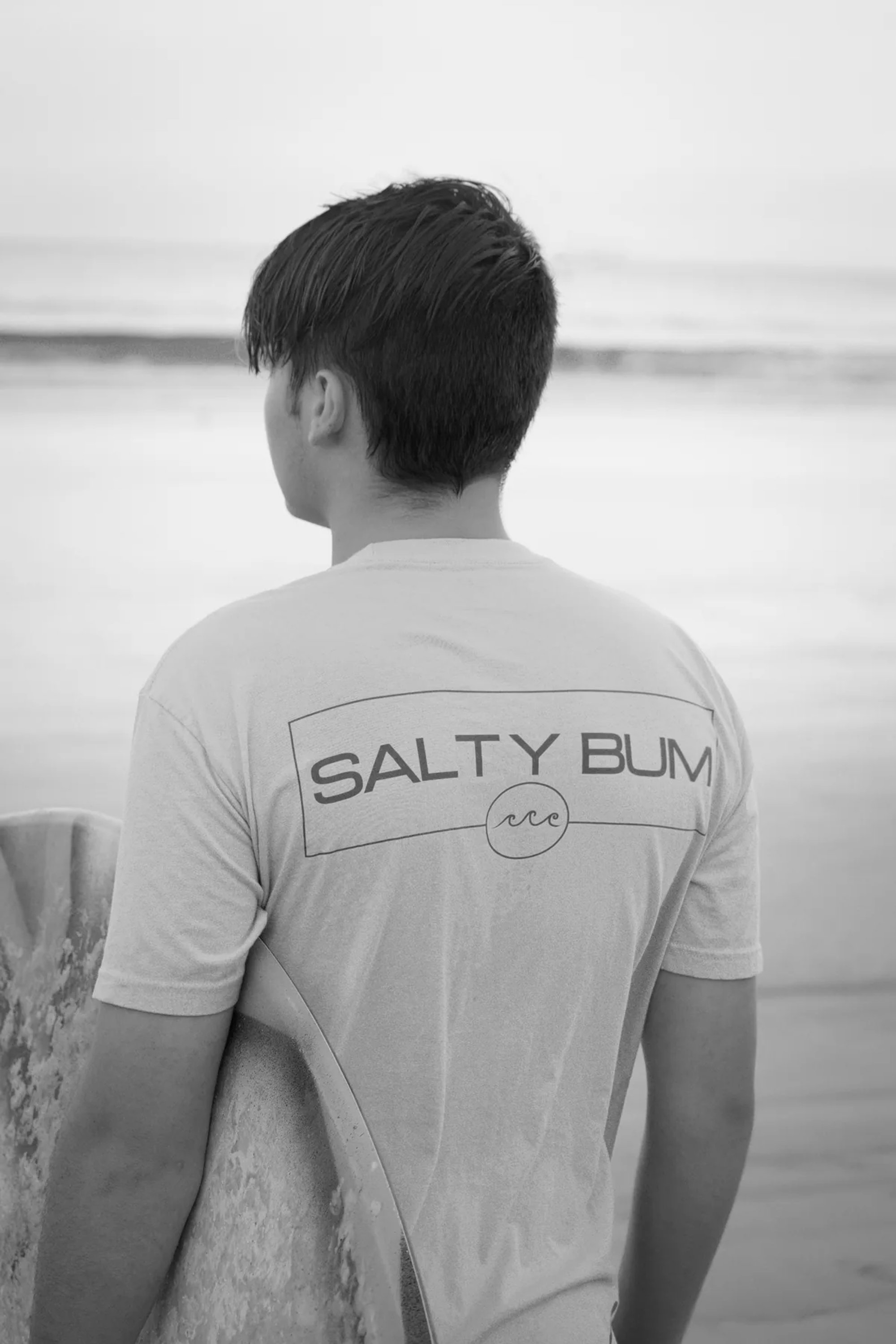 Man wearing a Salty Elm shirt while facing the ocean in black and white.