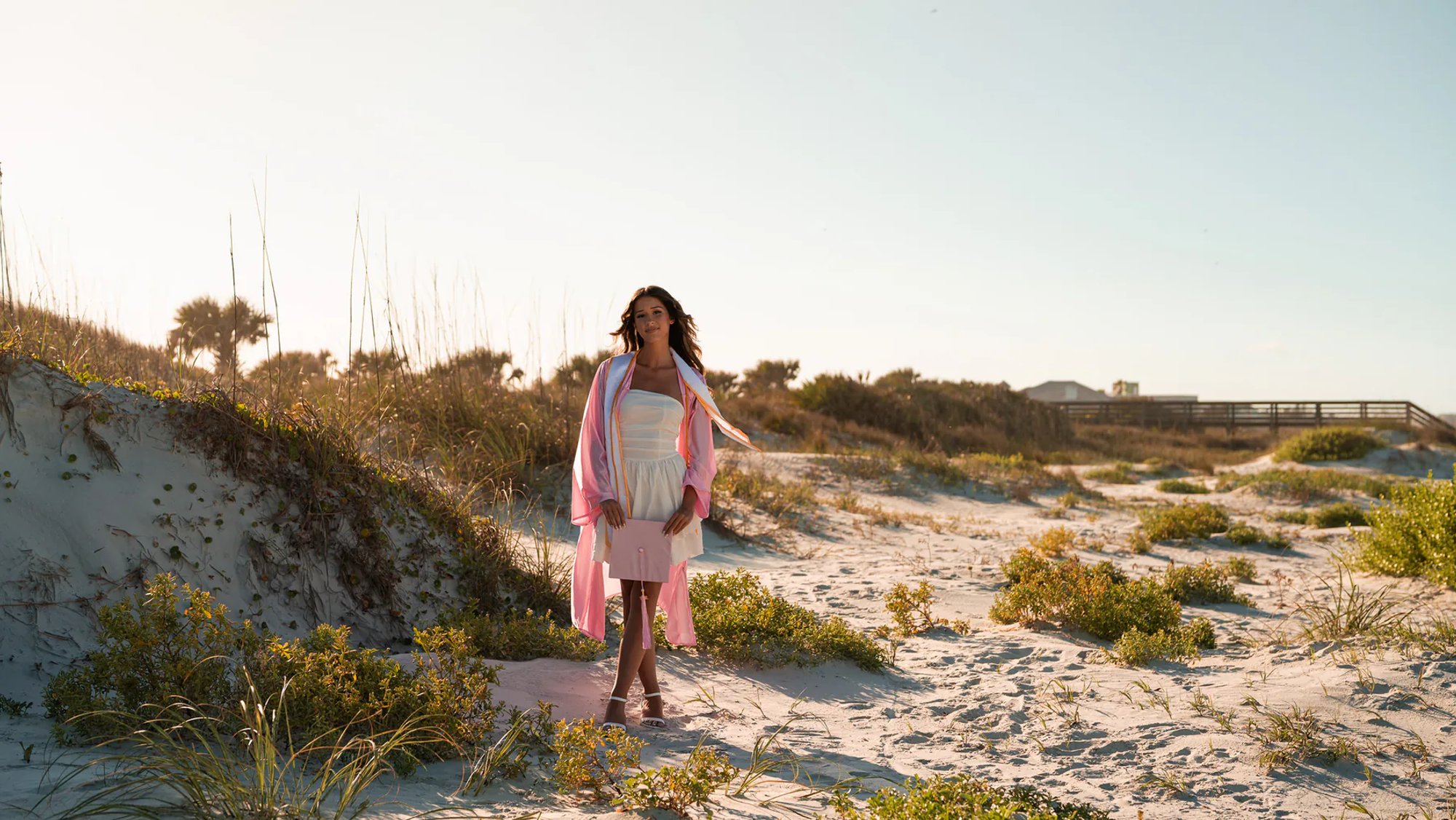 Young woman wearing a pink wrap over a white dress among coastal dunes.