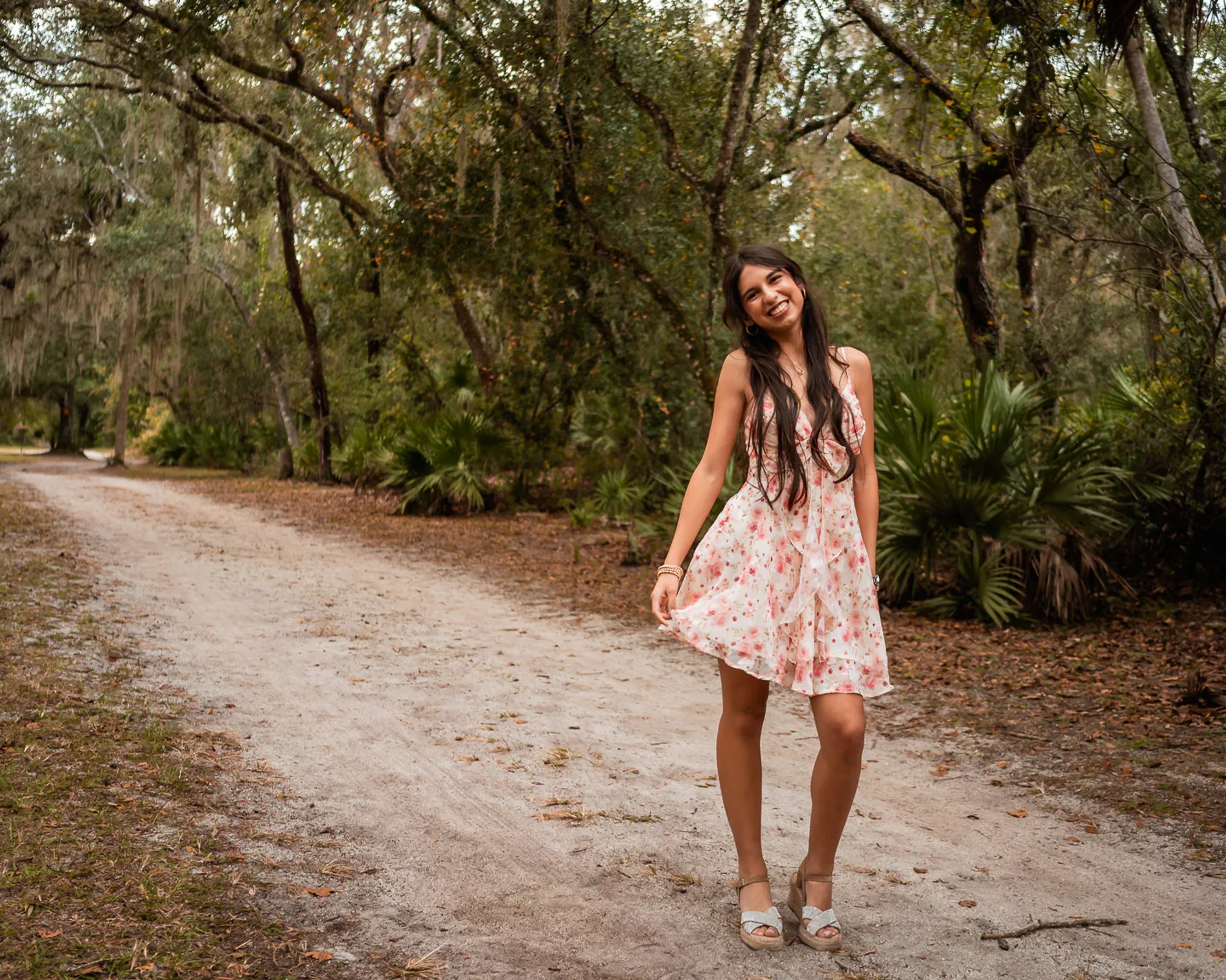 Young woman in a floral dress standing on a quiet wooded path.