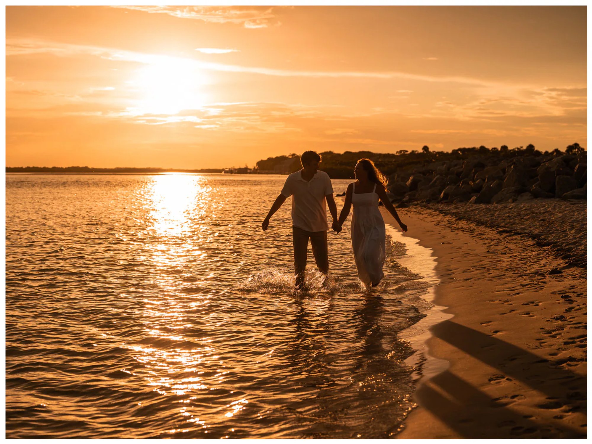 Couple walking hand in hand through shallow shoreline water.