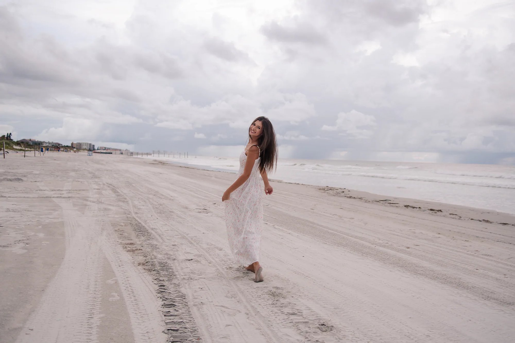 Daytona Beach senior portraits, Mia walking along the shoreline in a white floral dress, candid beach moment.