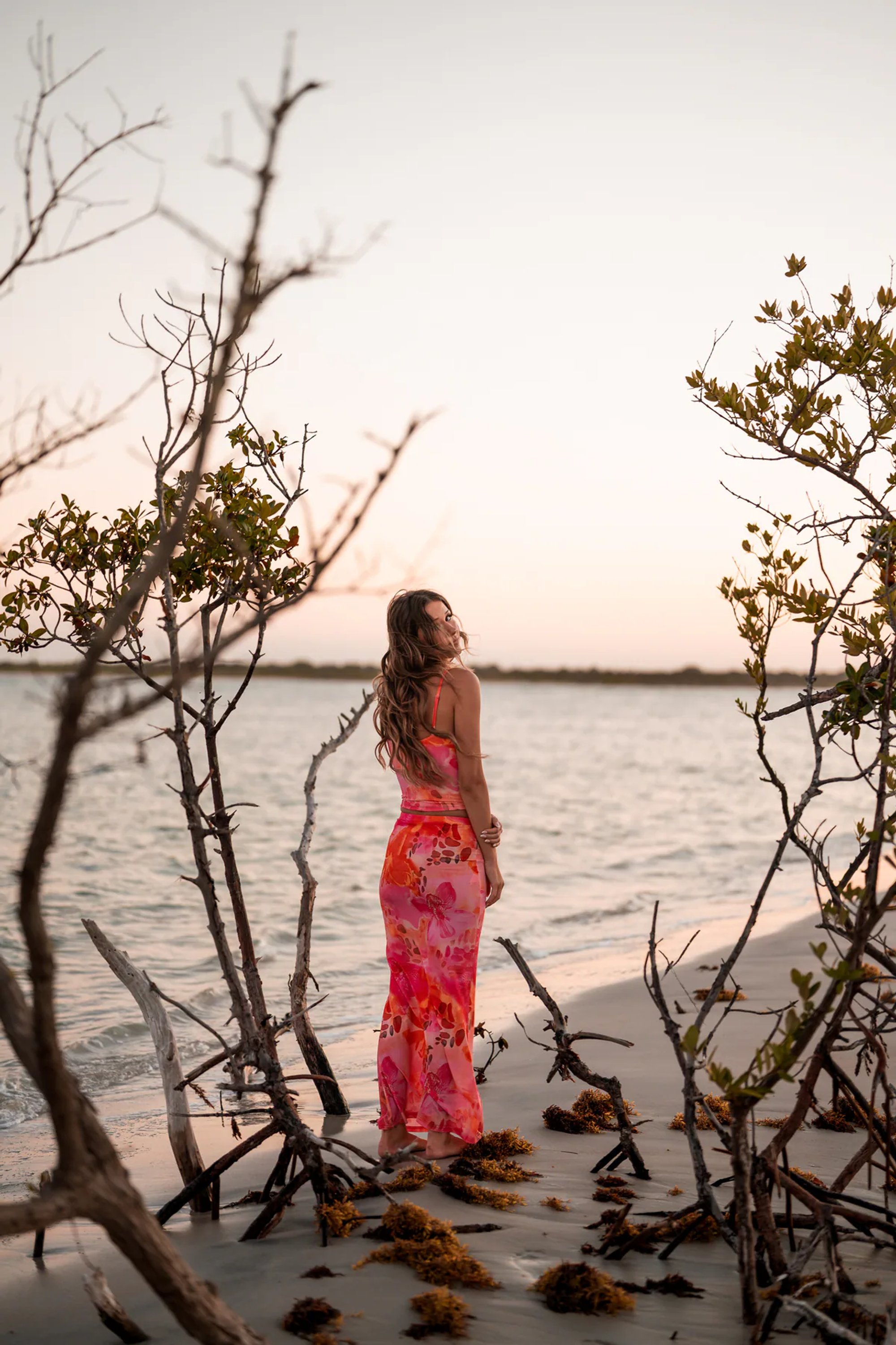 Young woman in a coral patterned dress standing among shoreline trees.