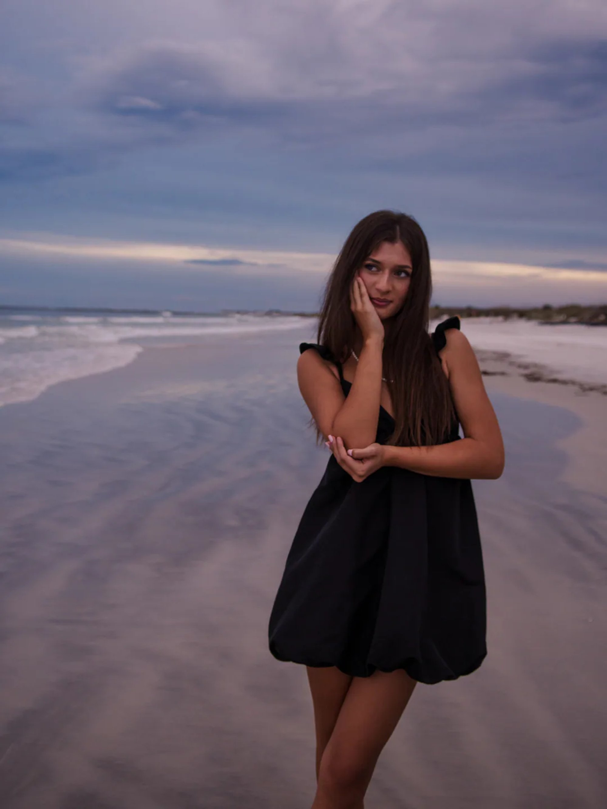 Dramatic senior portrait at Ponce Inlet Beach, Mia in a black dress standing in the sand with soft sunset skies.