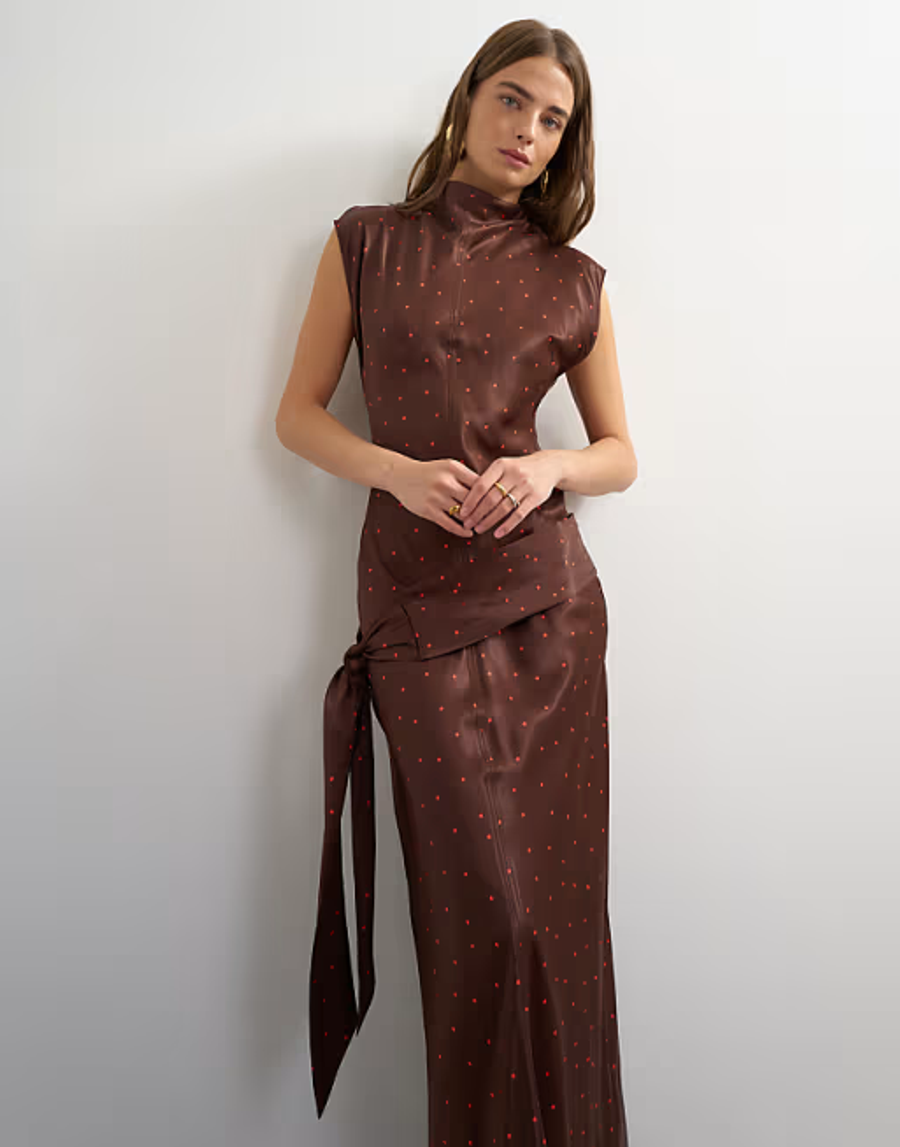 Model wearing a brown and red polka dot bias-cut satin dress in a studio setting with a concrete floor. The dress features a high neck with a slight cowl/fold at the neck with seam detailing at the centre front and centre back, which feature topstitch detailing on either side of the seam. At the dropped waist, features an Obi-style belt that goes asymmetrically across the dropped waist before tying at the wearer’s left-hand side. The back has a keyhole and button and loop closure at the neck to get into. The bias cut allows the fabric to fall naturally around the body.

Iole developed the pattern as the Creative Pattern Cutter, working through the bias-cut silhouette, fit and proportions, neckline. The fit and proportions were refined through toiling in CLO3D and draping on the stand, allowing the silhouette and construction to be checked and developed before finalising the pattern.