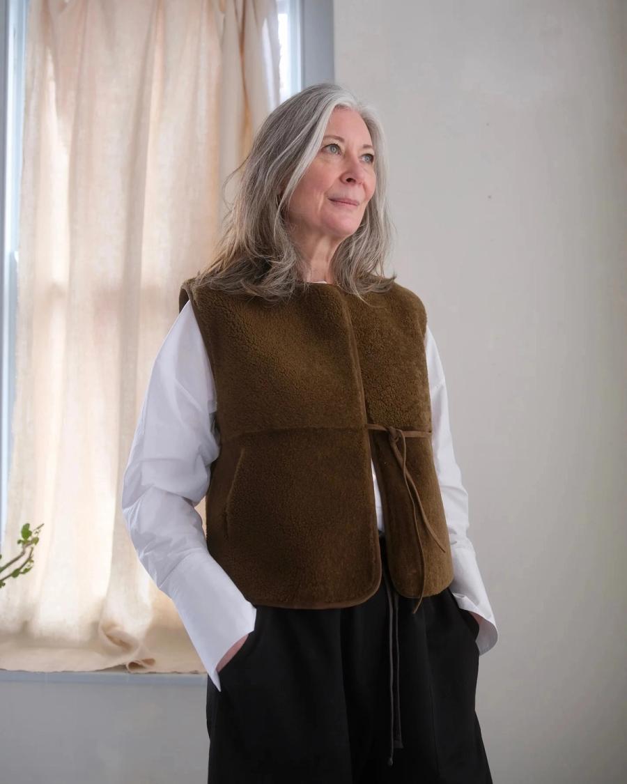Model wearing the reversible Camille Gilet by independent sustainable womenswear brand Studio Wylder, photographed in a home setting. Crafted from sheepskin sourced as a by-product of the food industry, the gilet features a wrap-over silhouette, round neckline and side-tie leather / suede fastenings. Reversible pockets and fully bound edges allow it to be worn on both sides, while the natural sheepskin construction gives the piece longevity and ensures it can be worn and passed down for years to come. Studio Iole worked as the freelance pattern cutter, developing the creative pattern through fittings and refinements and working closely on the fit, silhouette and reversible construction through to the final production pattern used to create the finished garment shown.